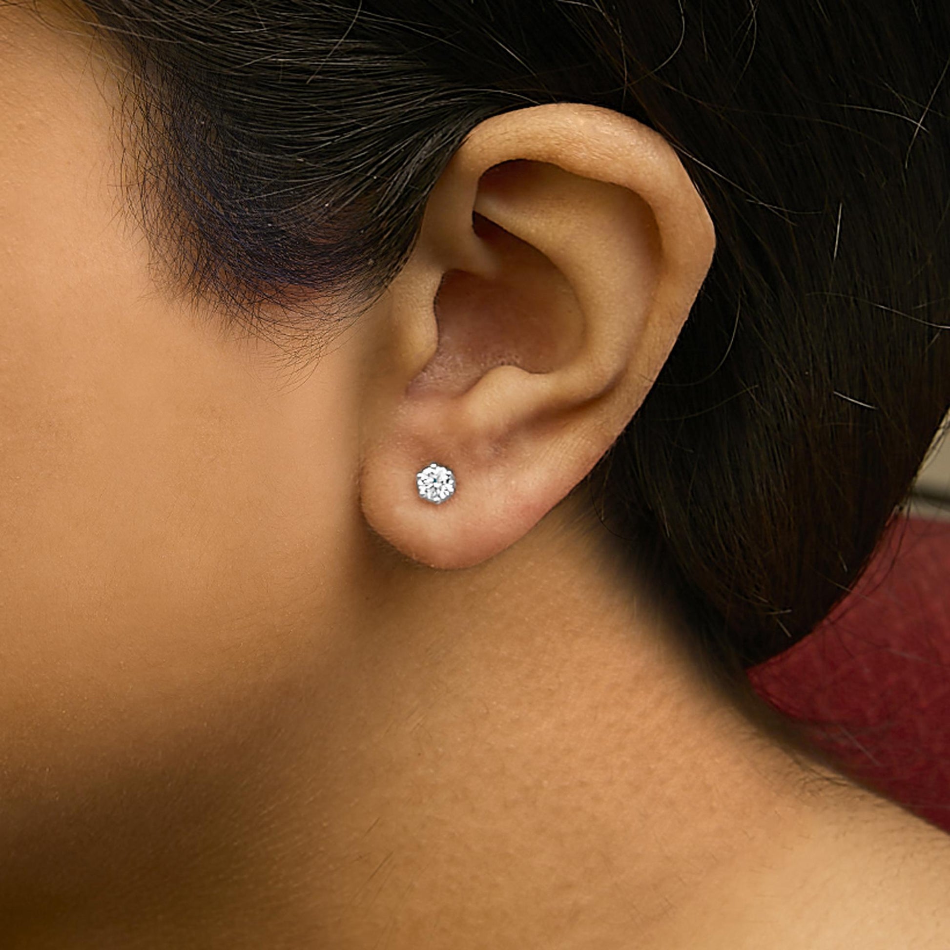 Close-up side view of a model wearing a white metal stud earring with a single round white gemstone in a six-prong setting.