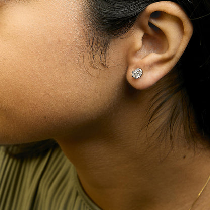 Close-up side view of a woman's face wearing white metal stud earrings with two round prong-set white gemstones surrounded by multiple smaller round pave-set white gemstones.