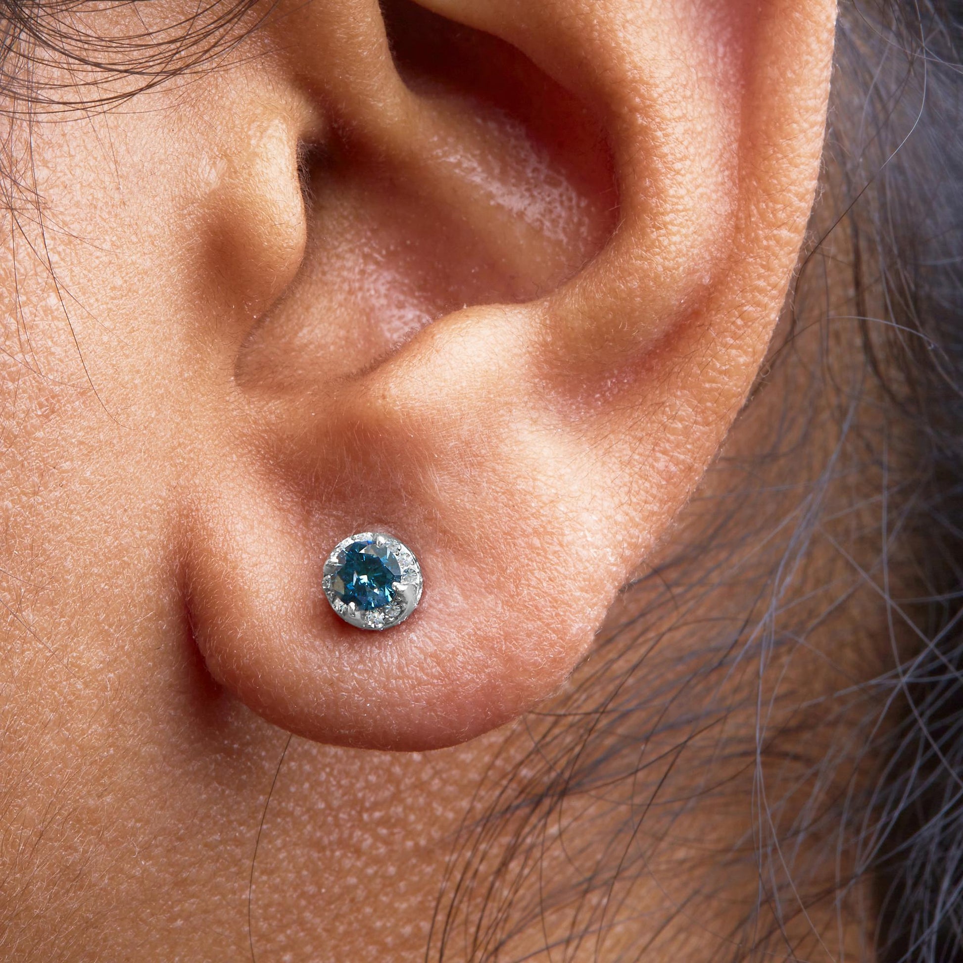 Close-up side view of a white metal earring worn on a model's ear, featuring a round blue gemstone in prong setting surrounded by small round white gemstones in pave setting.
