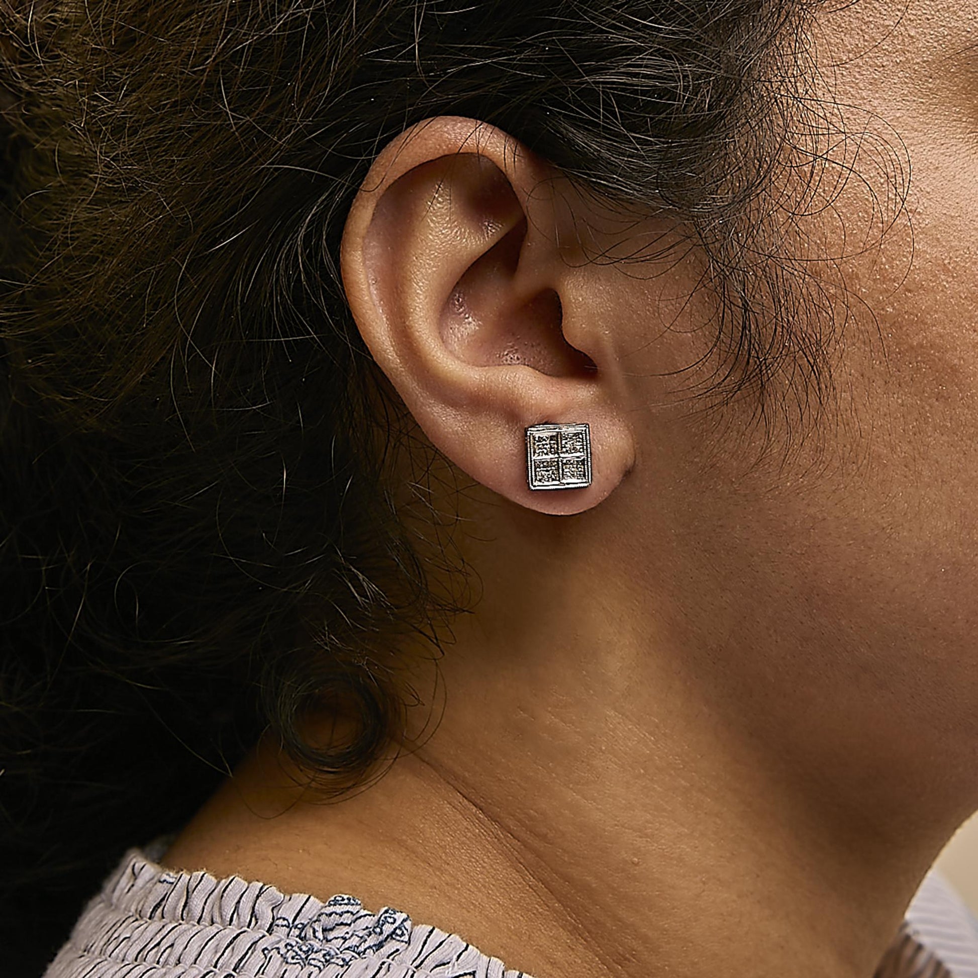 Close-up side view of a white metal square-shaped stud earring with multiple small white gemstones in an invisible setting, worn on a model's ear.