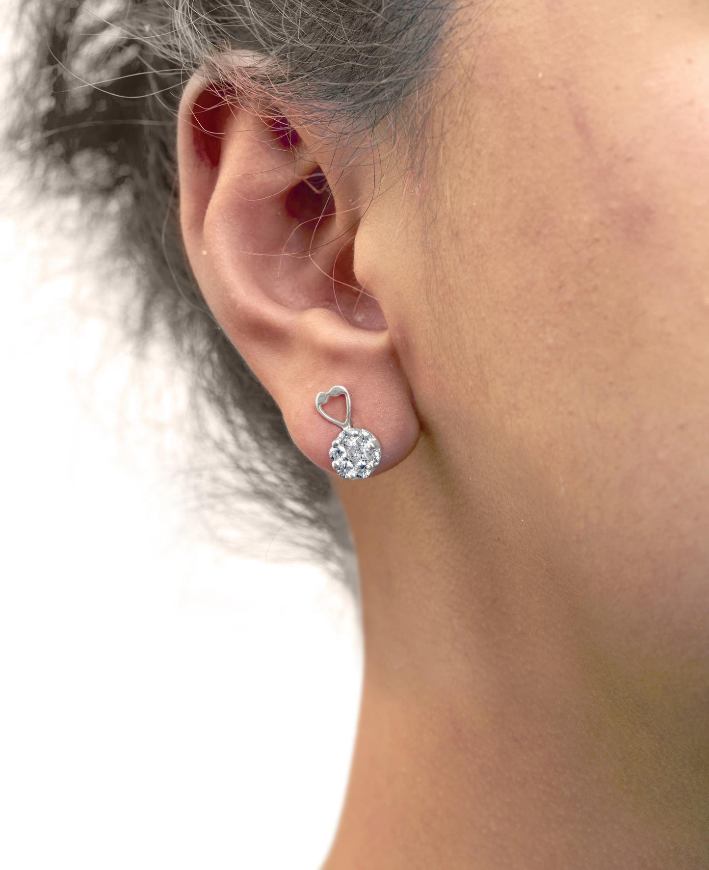 Side view of white metal earrings with a heart-shaped detail and clustered round white gemstones, worn on a model's ear.