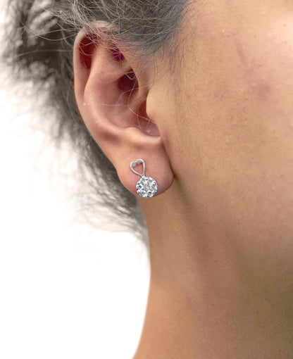 Side view of white metal earrings with a heart-shaped detail and clustered round white gemstones, worn on a model's ear.
