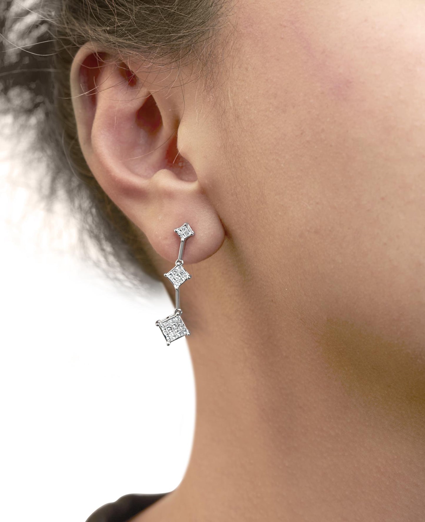 Close-up side view of a model wearing white metal drop earrings with three connected princess-shaped sections featuring clusters of white gemstones in an invisible setting.