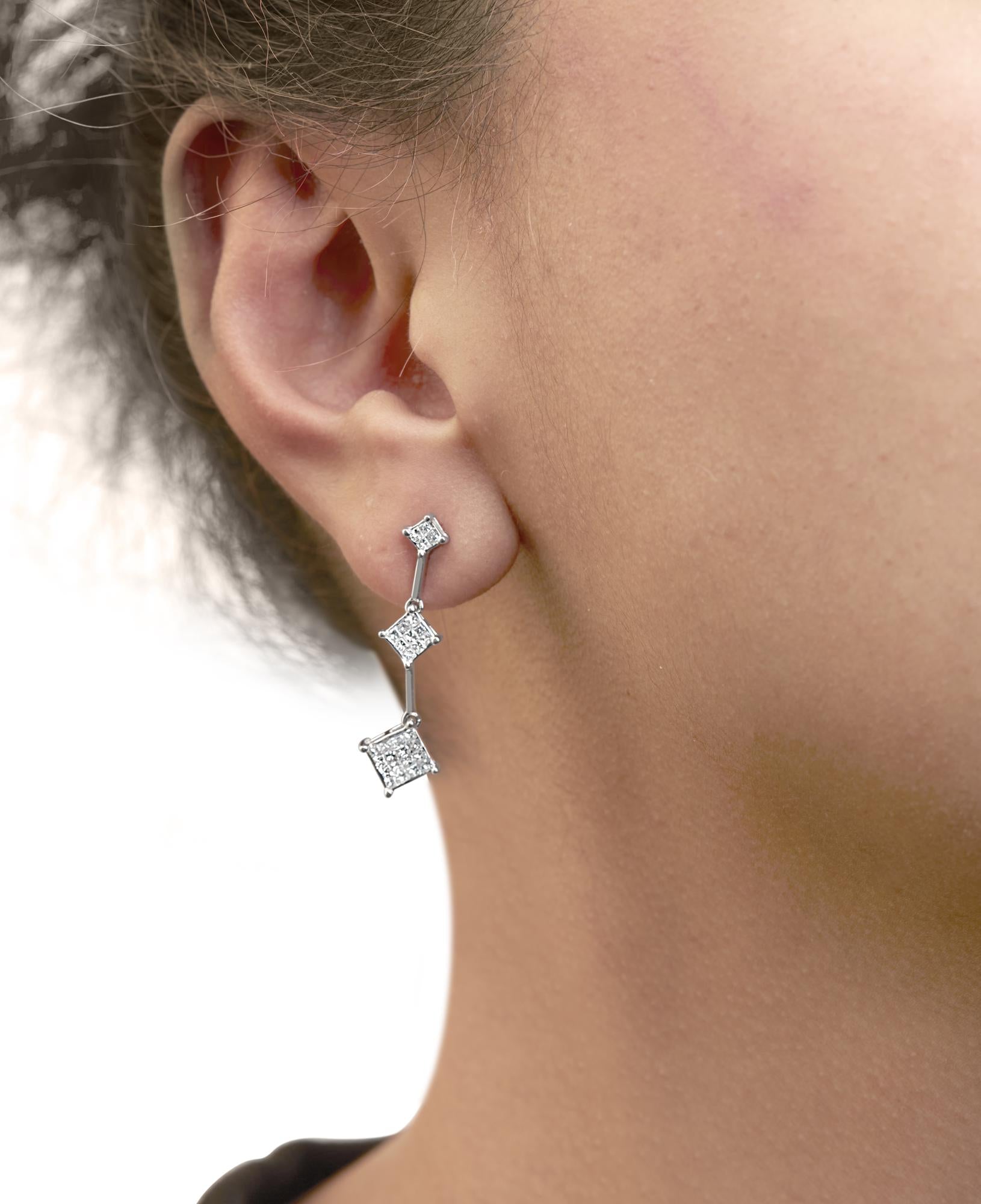 Close-up side view of a model wearing white metal drop earrings with three connected princess-shaped sections featuring clusters of white gemstones in an invisible setting.