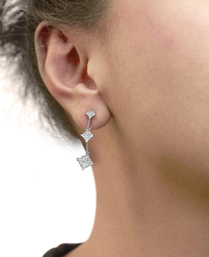 Close-up side view of a model wearing white metal drop earrings with three connected princess-shaped sections featuring clusters of white gemstones in an invisible setting.