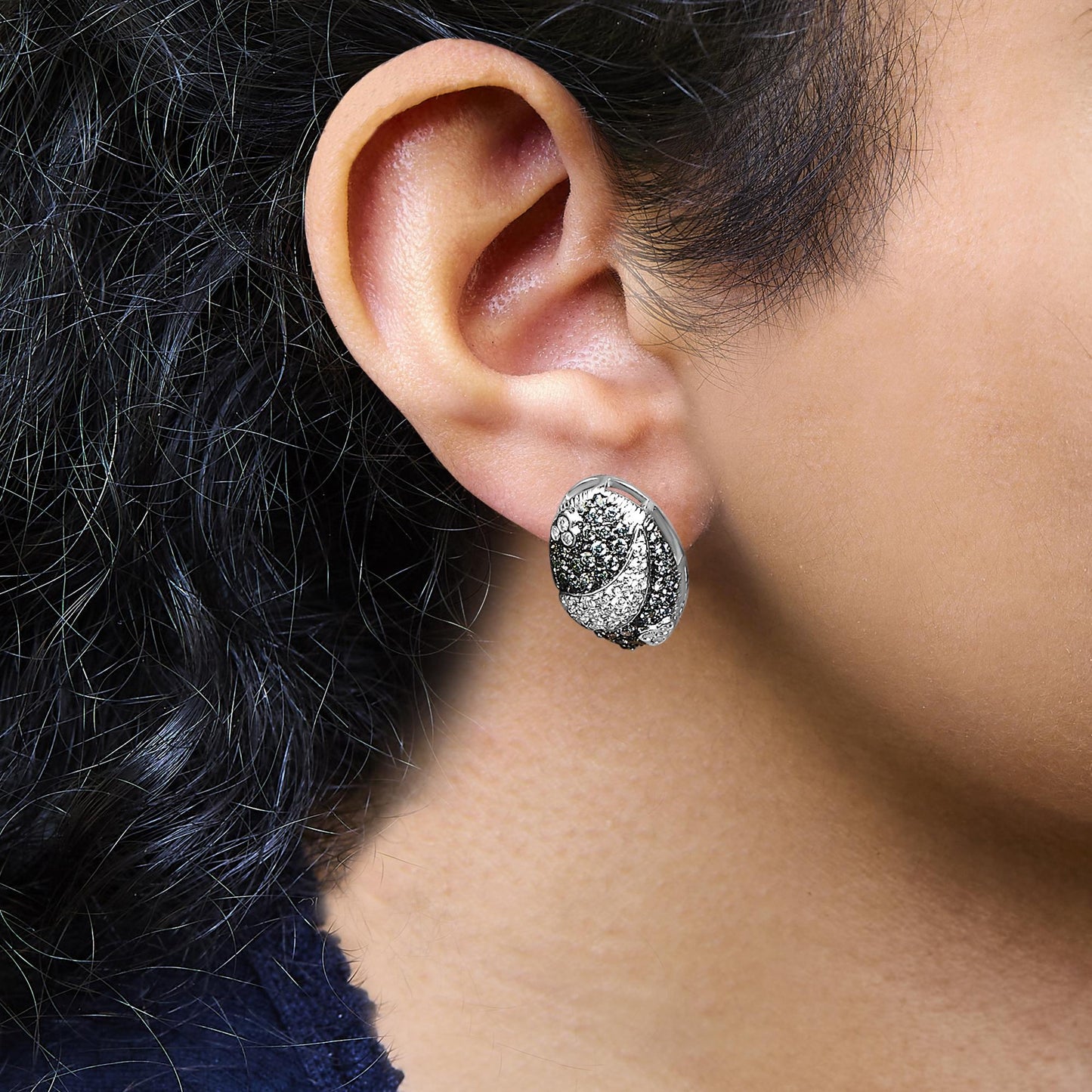White metal oval-shaped earrings with pave-set round blue and white gemstones, shown worn on a model, close-up side view.