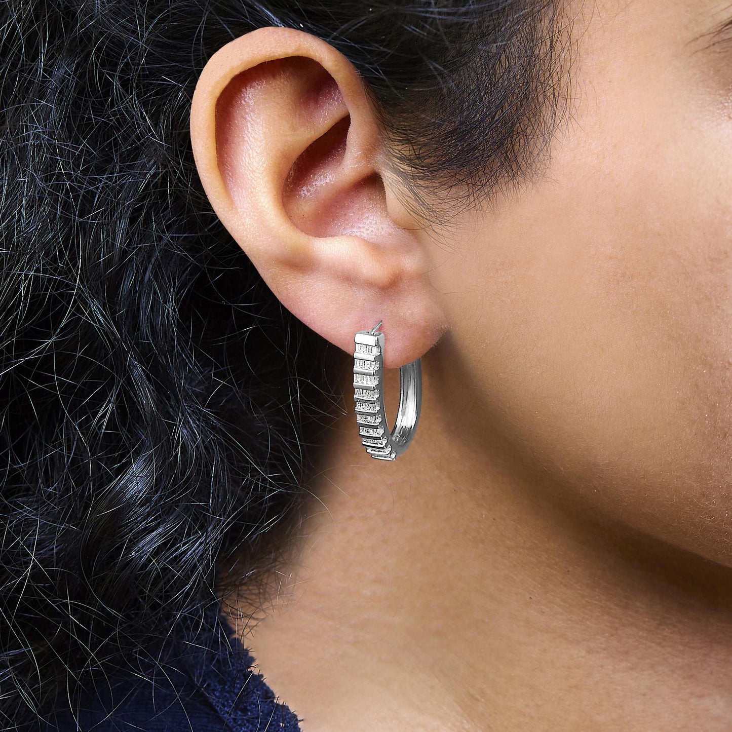 Close-up side view of a white metal hoop earring on a model's ear, featuring a row of white baguette-shaped gemstones set in a channel setting along the front.