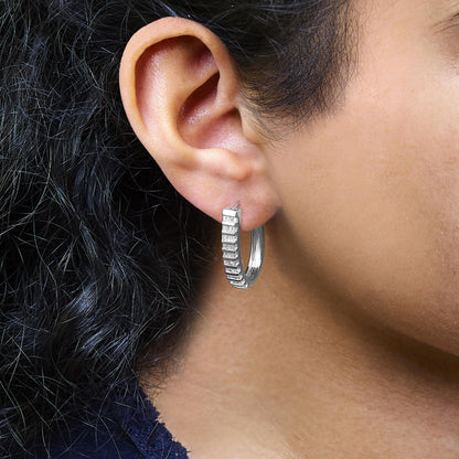 Close-up side view of a white metal hoop earring on a model's ear, featuring a row of white baguette-shaped gemstones set in a channel setting along the front.