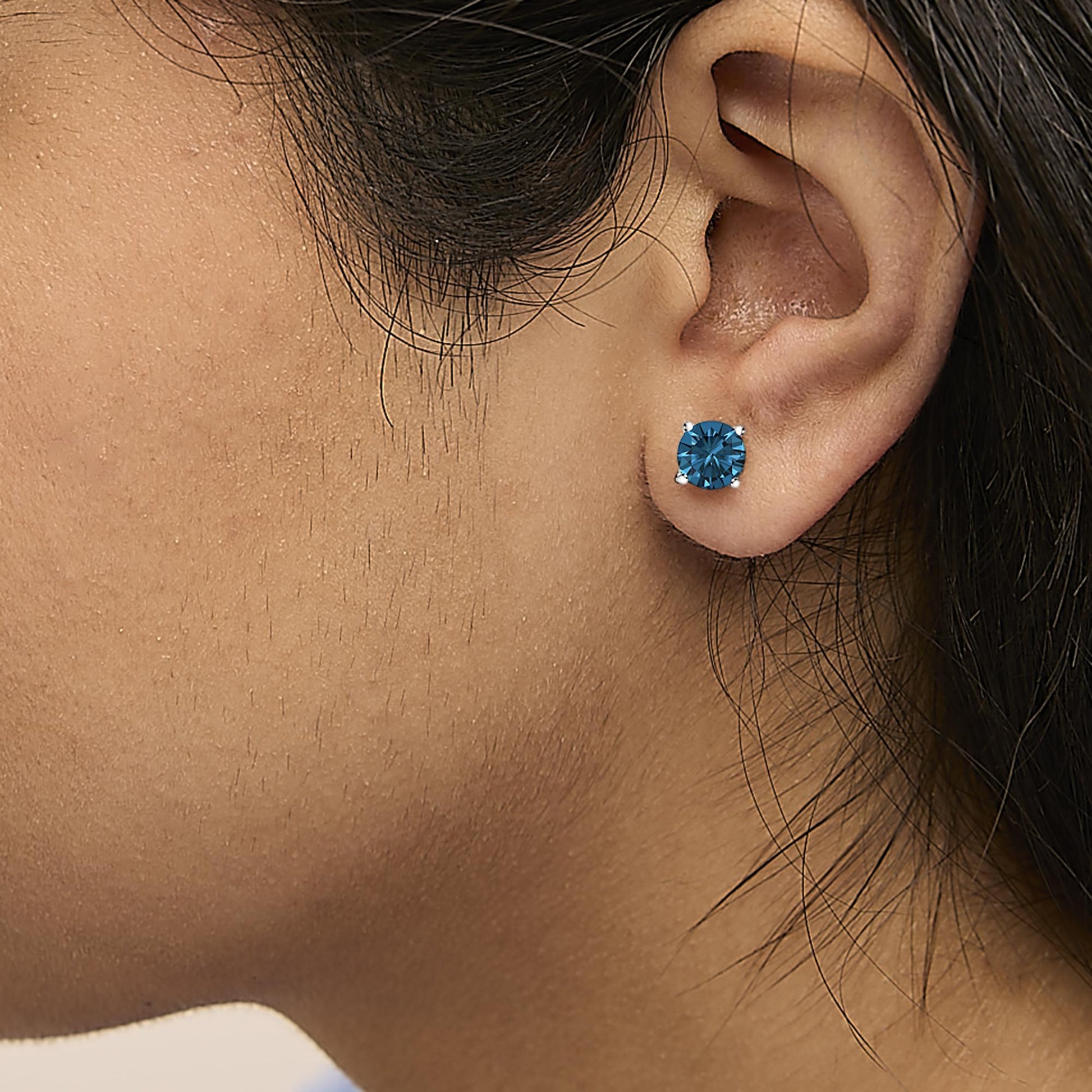 Close-up side view of a white metal stud earring with a round blue gemstone in a four-prong setting, worn on a model's ear.