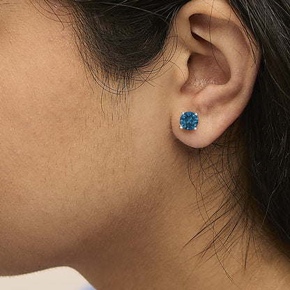 Close-up side view of a white metal stud earring with a round blue gemstone in a four-prong setting, worn on a model's ear.