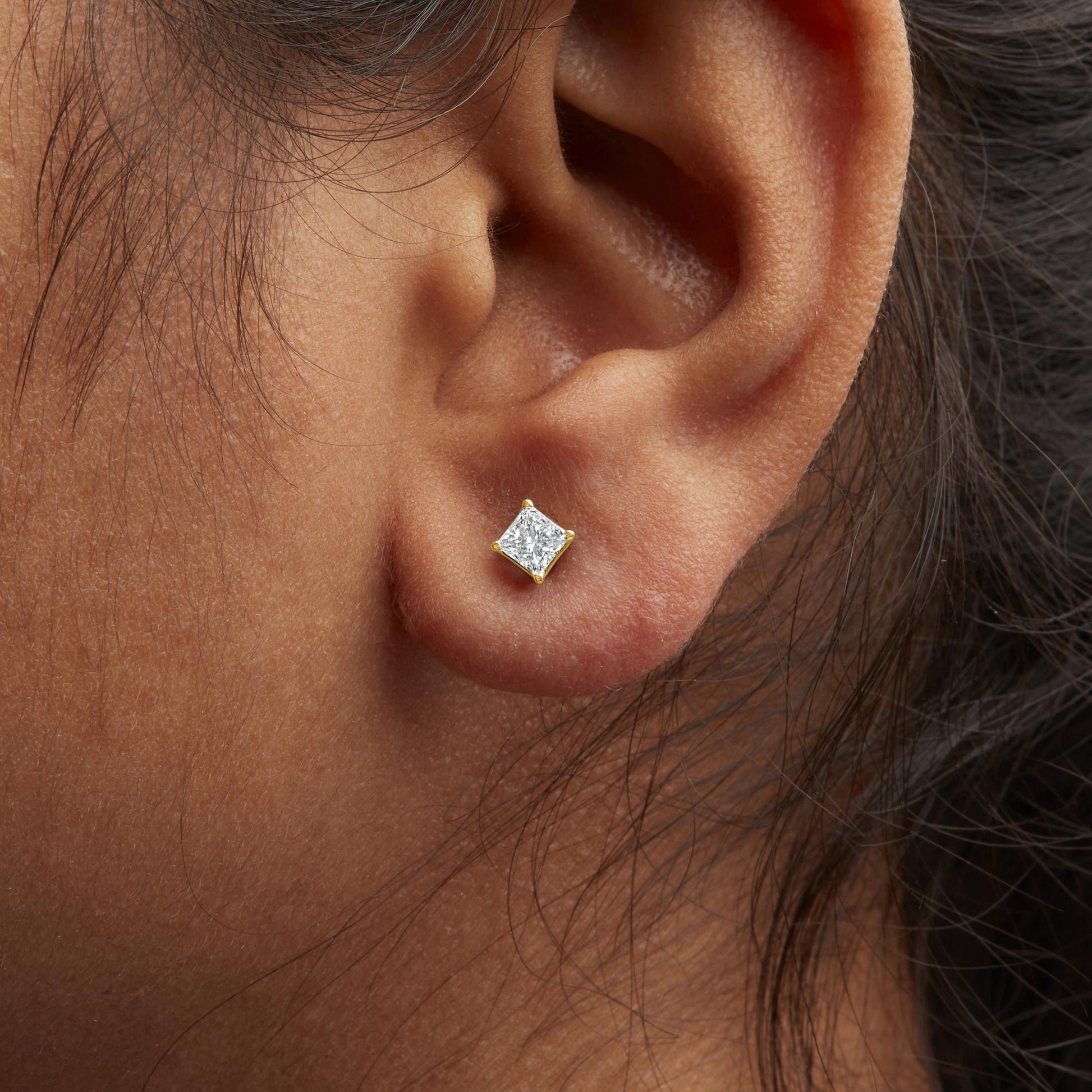 Close-up side view of a yellow metal earring with a princess-shaped white gemstone in a four-prong setting worn on a model's ear.