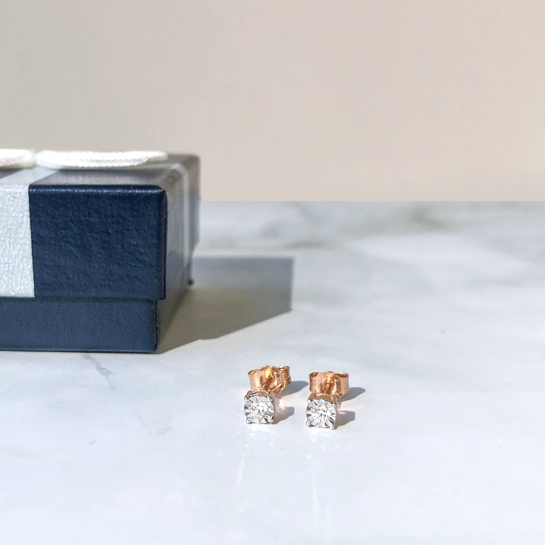 Pair of rose-colored stud earrings with round white gemstones in a miracle setting, shown at a slight angle on a marble surface with a navy and silver gift box in the background.