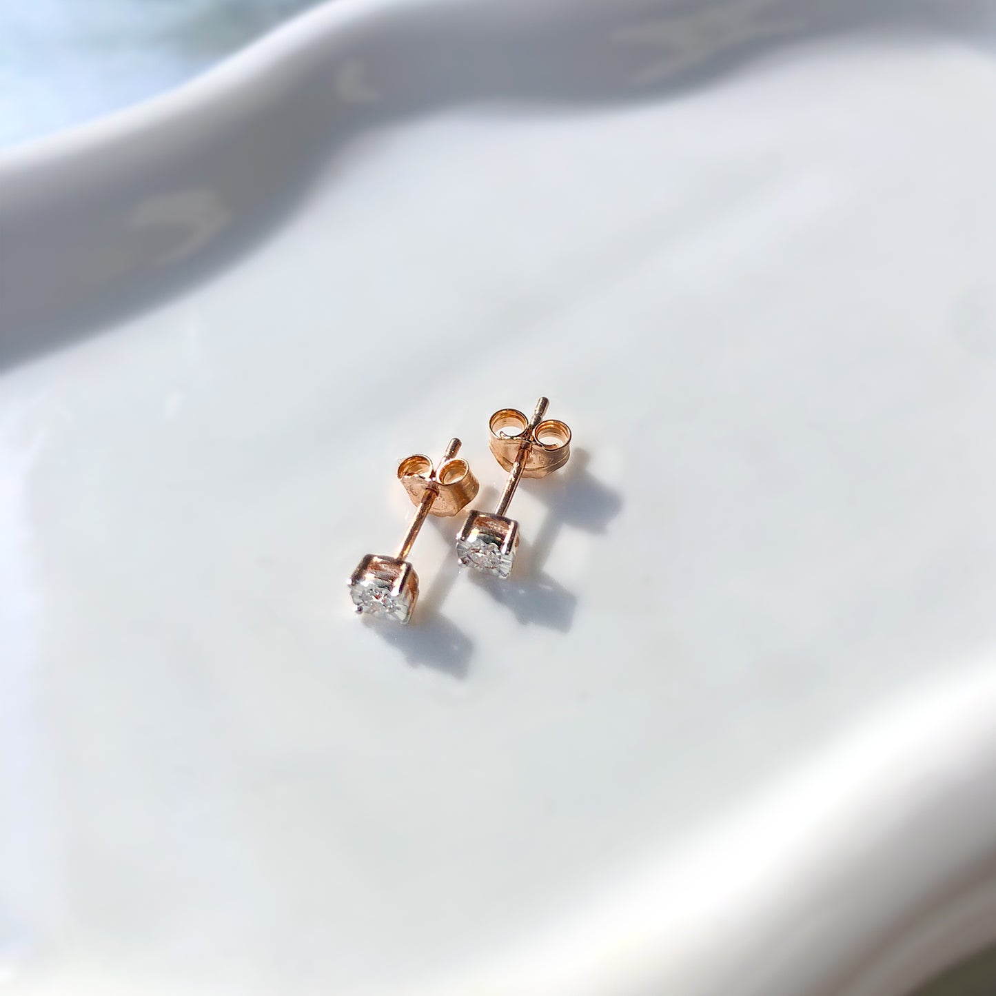Pair of rose-colored stud earrings with round white gemstones in a miracle setting, shown in a close-up angled shot on a smooth white surface.