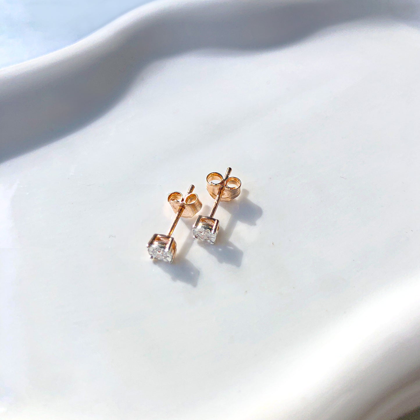 Pair of rose-colored stud earrings with round white gemstones in a miracle setting, shown from a top angle on a white textured surface.