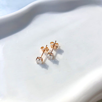 Pair of rose-colored stud earrings with round white gemstones in a miracle setting, shown from a top angle on a white textured surface.