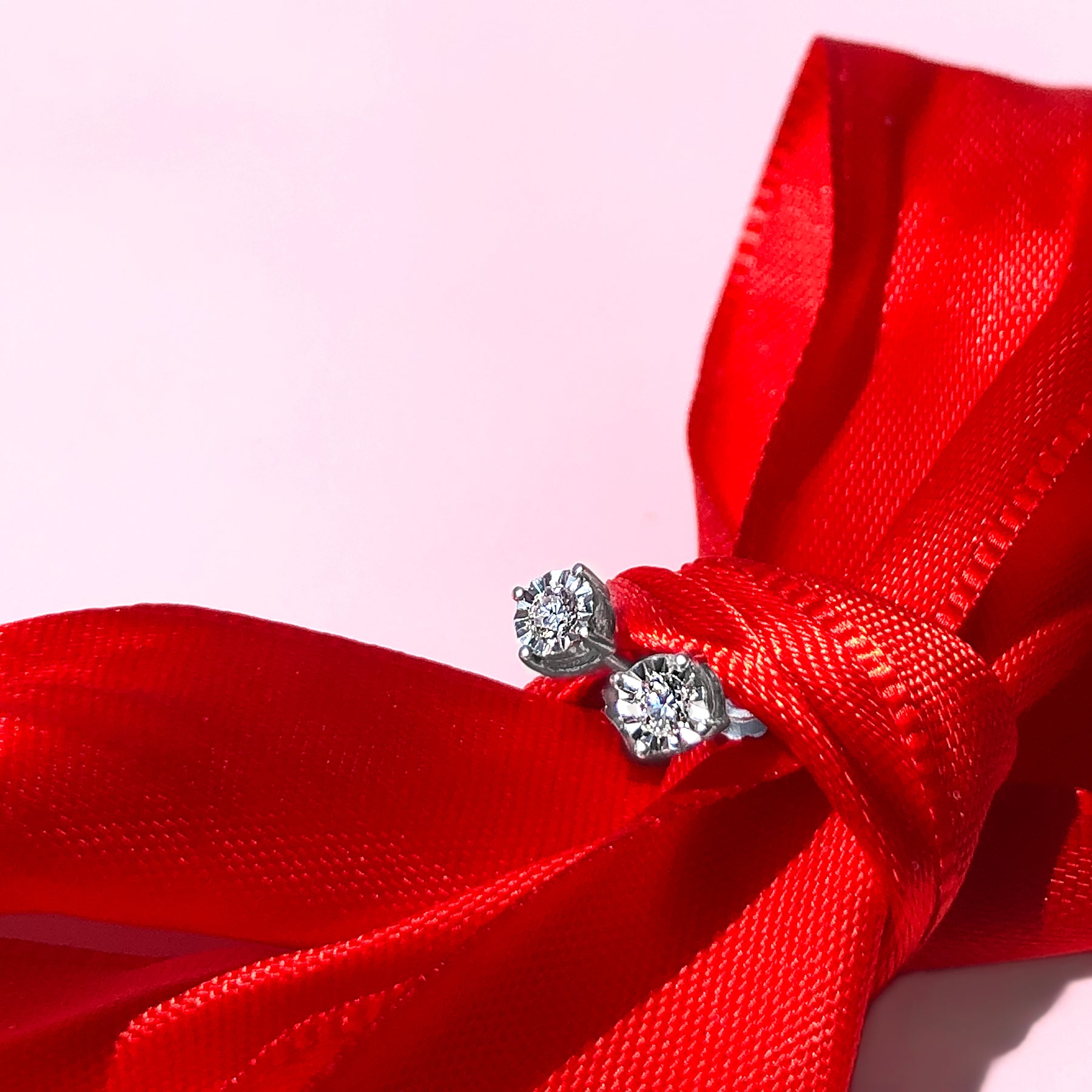 Close-up angled shot of a pair of white metal stud earrings with round white gemstones in miracle settings, displayed tied on a red ribbon.