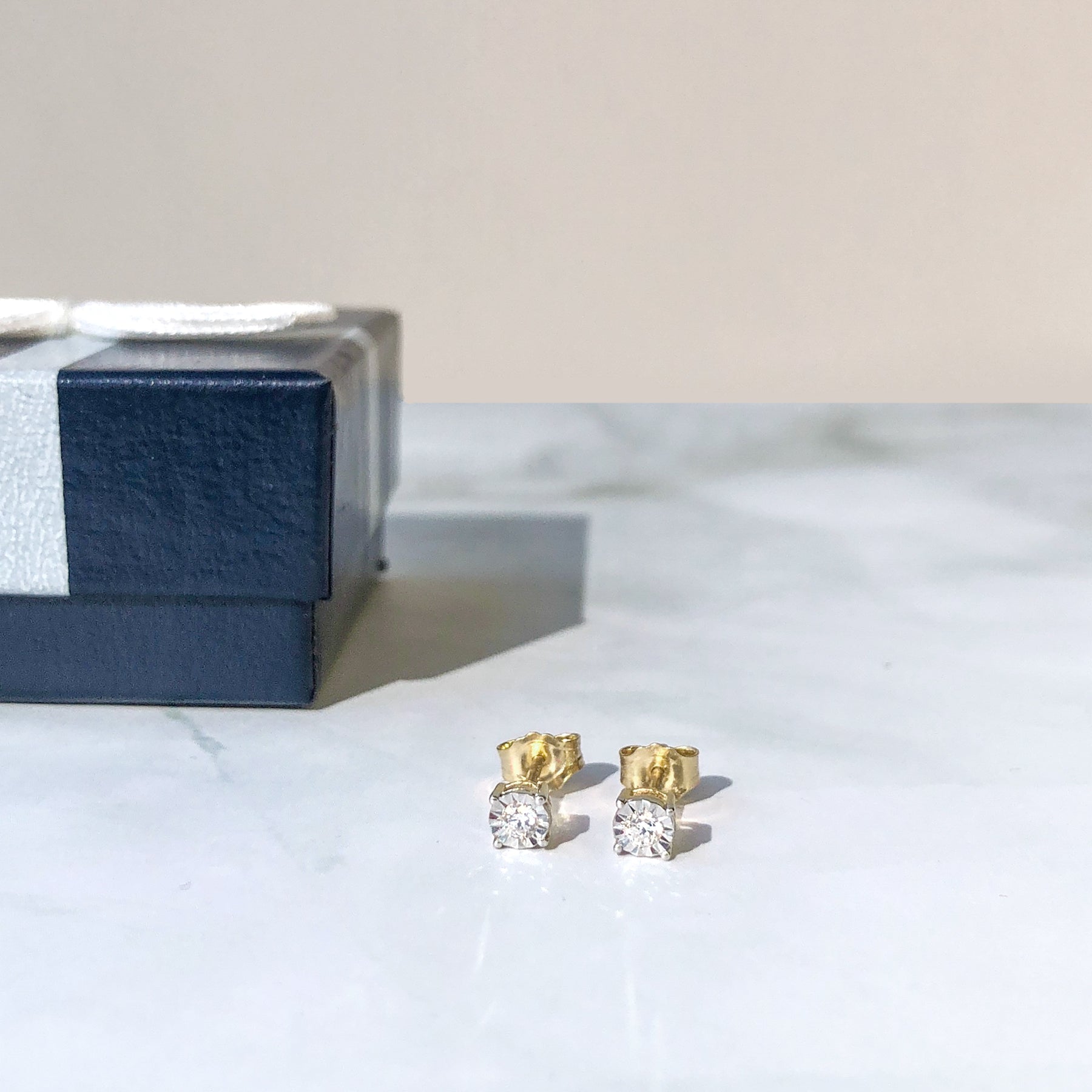 Pair of yellow metal stud earrings with round white gemstones in miracle settings, shown from front on a white surface next to a dark blue gift box.