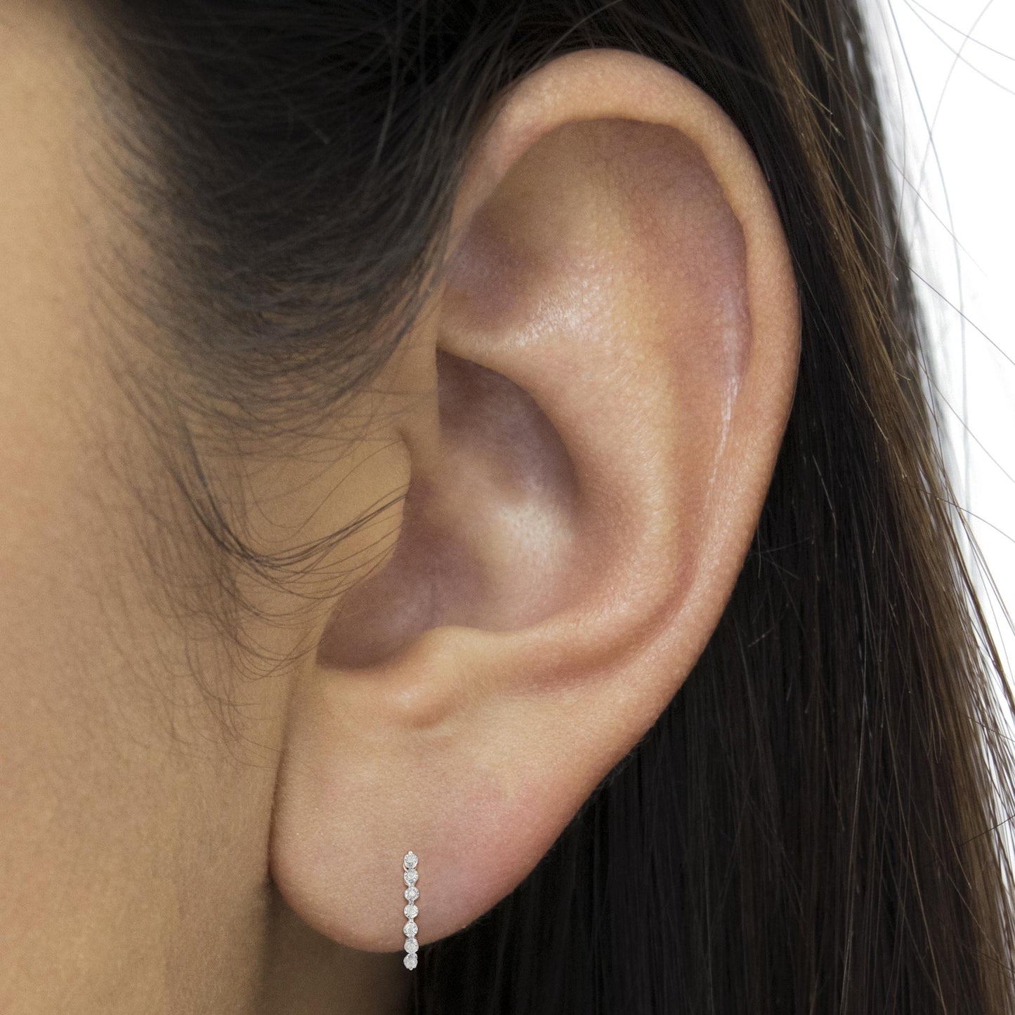 Close-up side view of an ear on a model wearing a white metal earring with a vertical row of small round white gemstones in a miracle setting.