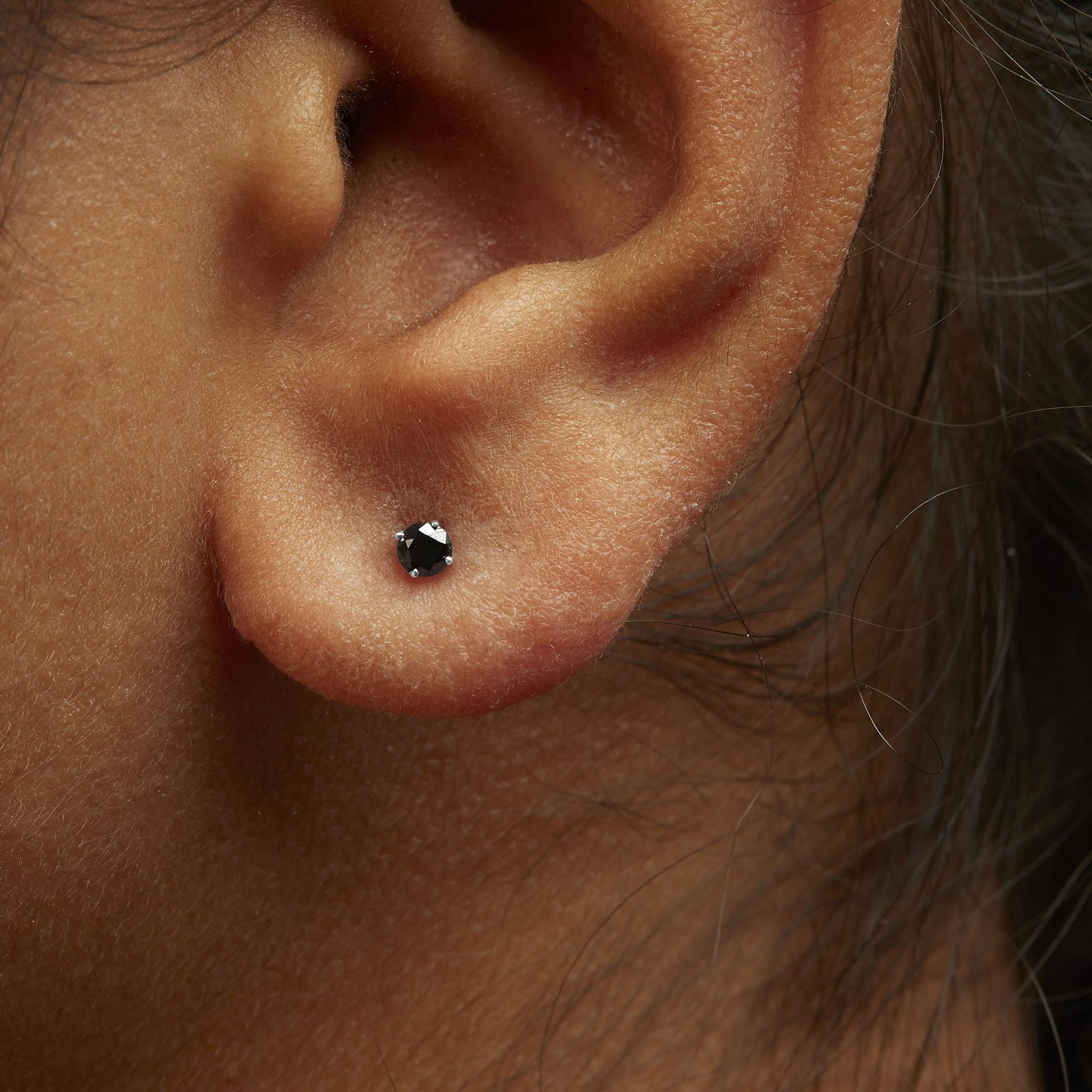 Close-up side angle of an ear wearing a white metal stud earring with a round black gemstone in prong setting.