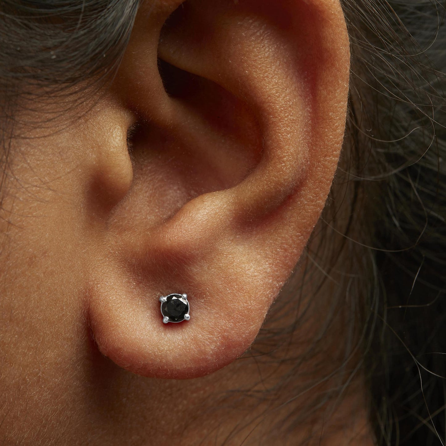 Close-up side view of a white metal stud earring with a round black gemstone in a 4-prong setting, worn on a model's ear.