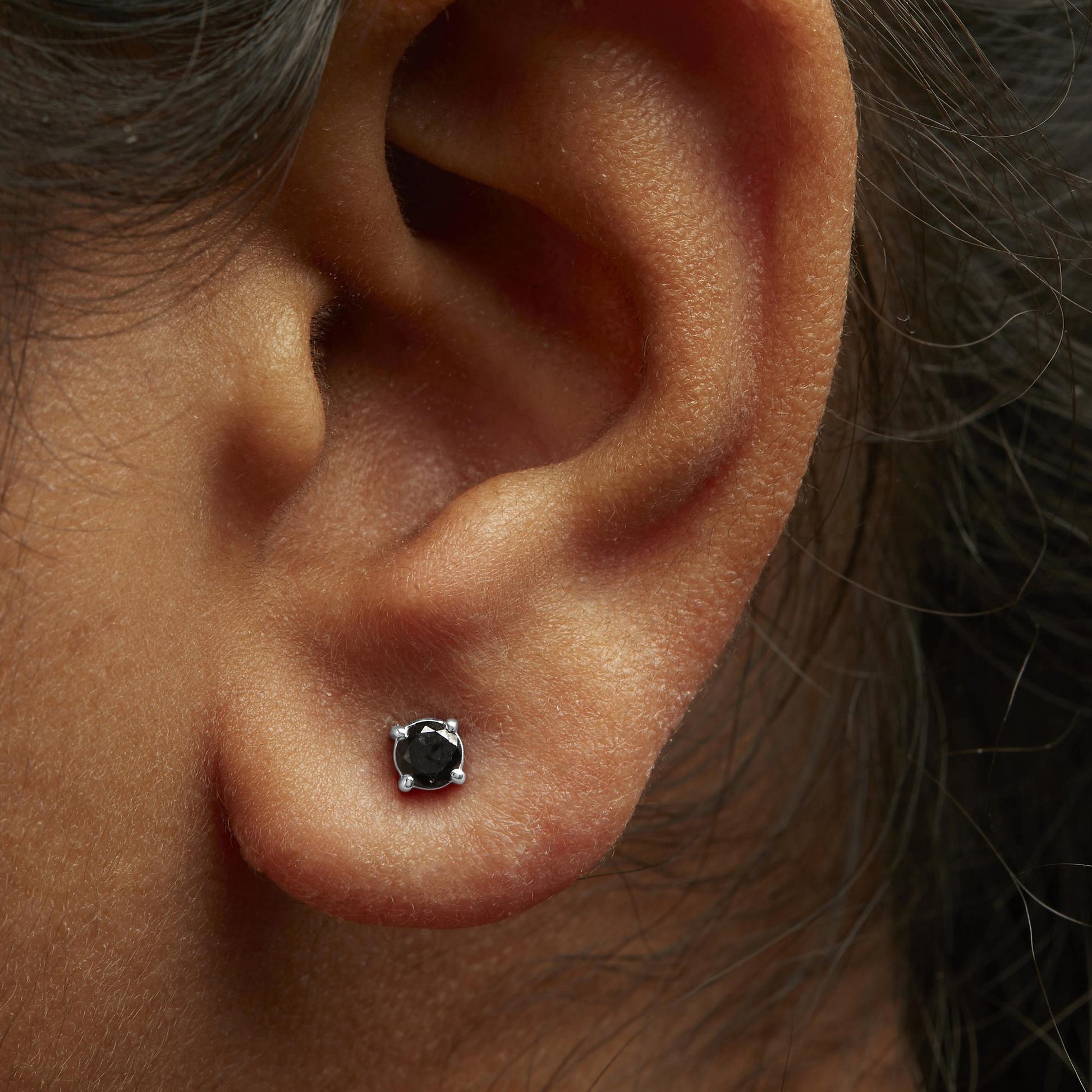 Close-up side view of a white metal stud earring with a round black gemstone in a 4-prong setting, worn on a model's ear.