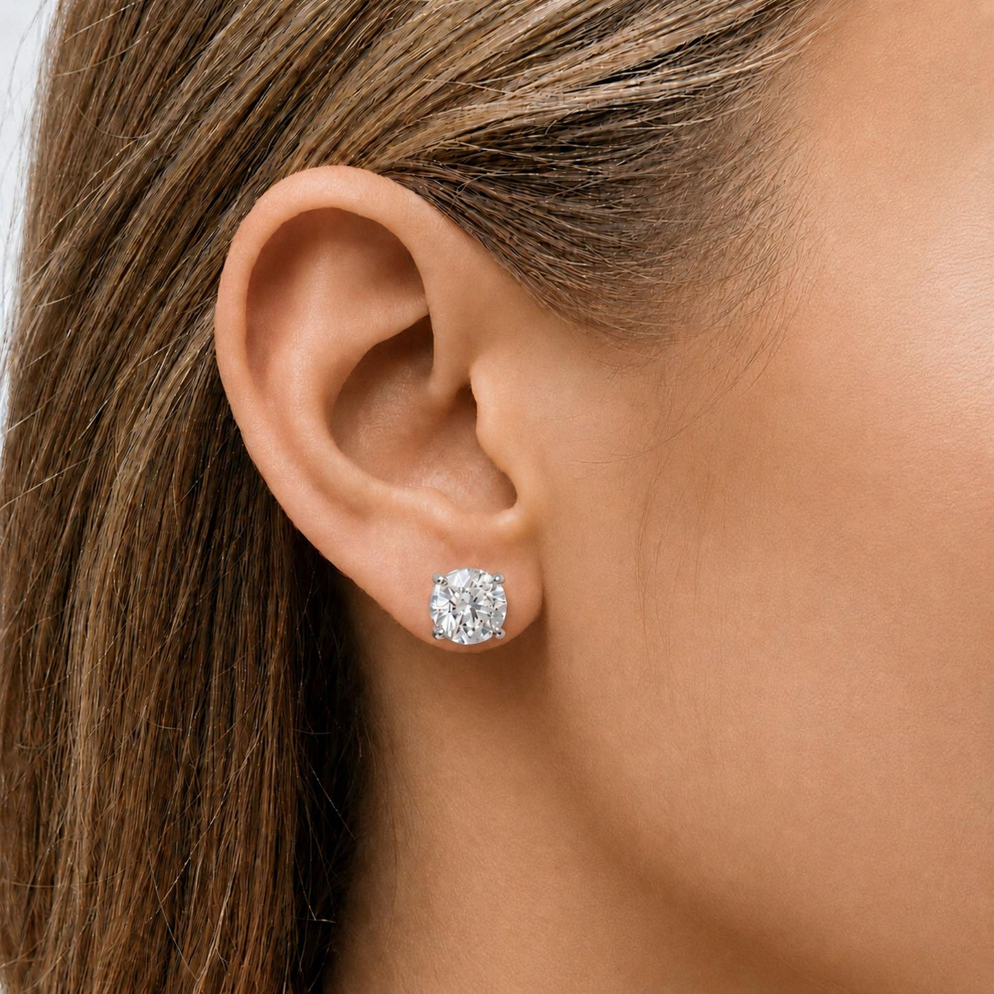 Close-up side view of a model wearing a pair of white metal stud earrings, each featuring a round white gemstone set in a four-prong setting.
