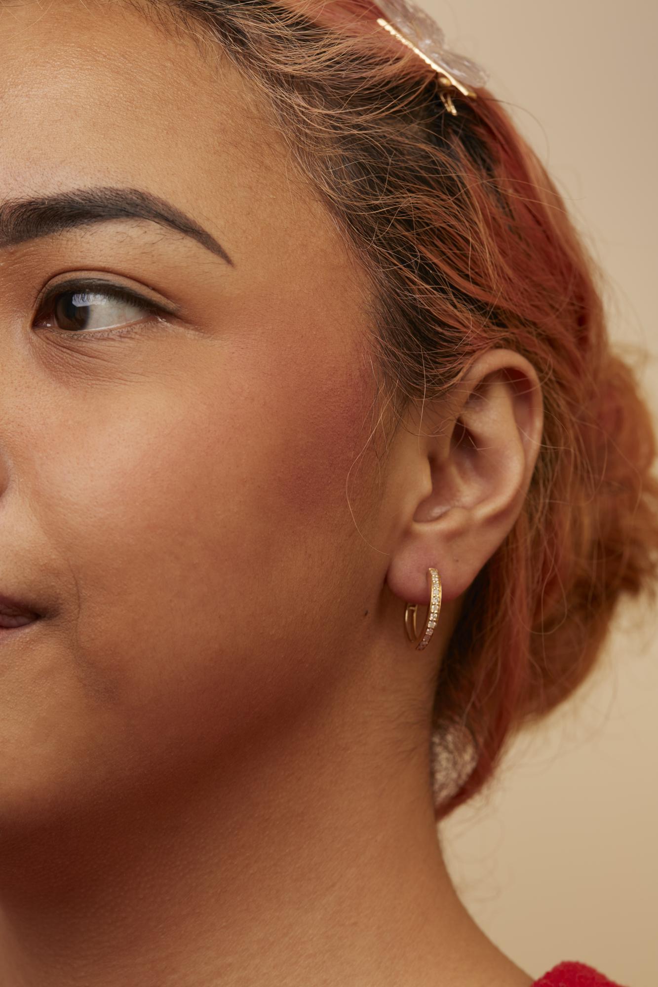 Close-up side angle of a model wearing yellow metal hoop earrings set with multiple white round gemstones in prong settings.