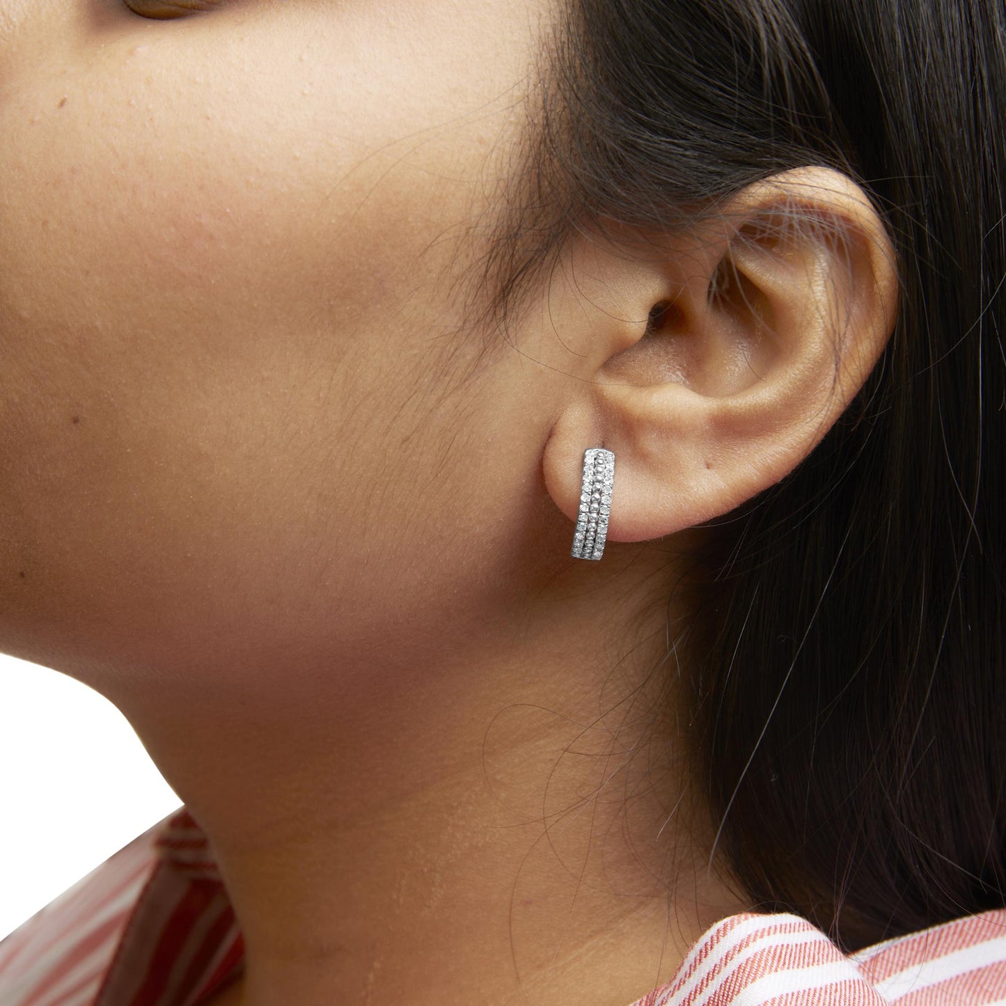 Close-up side view of a white metal earring with three rows of round white gemstones in pave setting, worn on a model's ear.