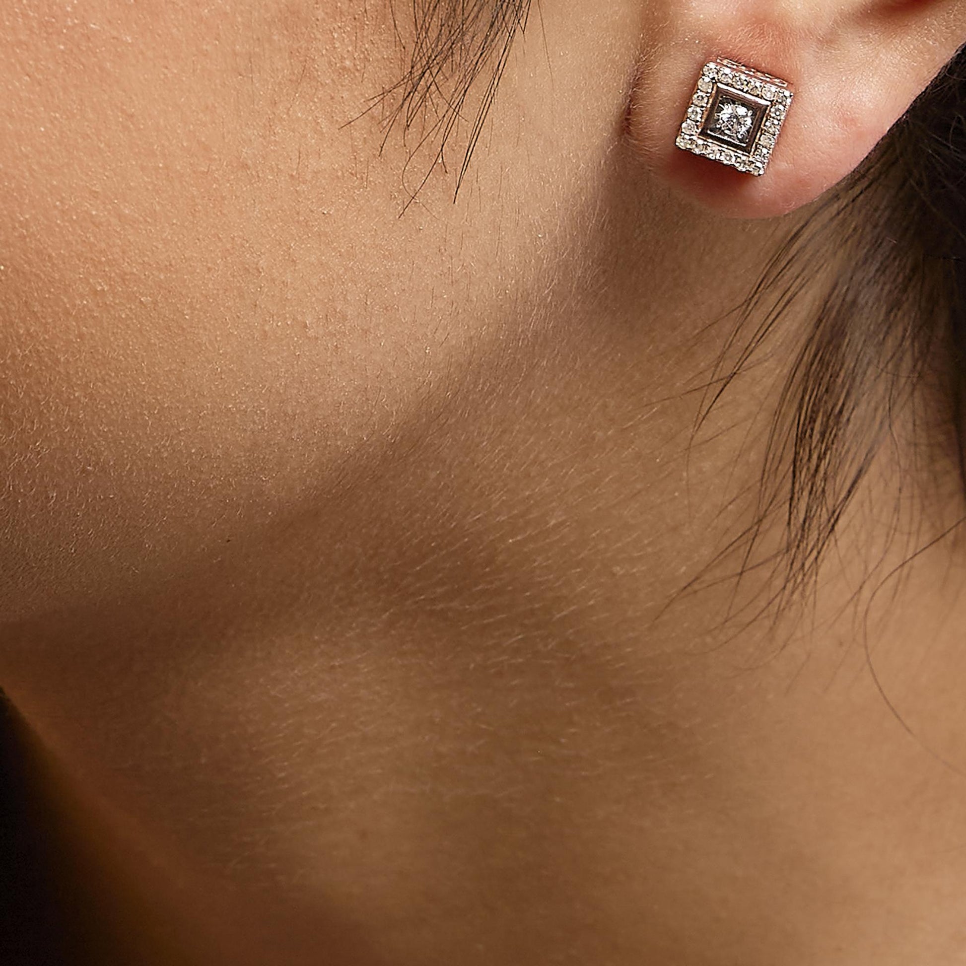 Close-up side view of a rose metal square-shaped stud earring on a model’s ear, featuring a central white gemstone in a miracle setting surrounded by multiple small white gemstones in prong settings.