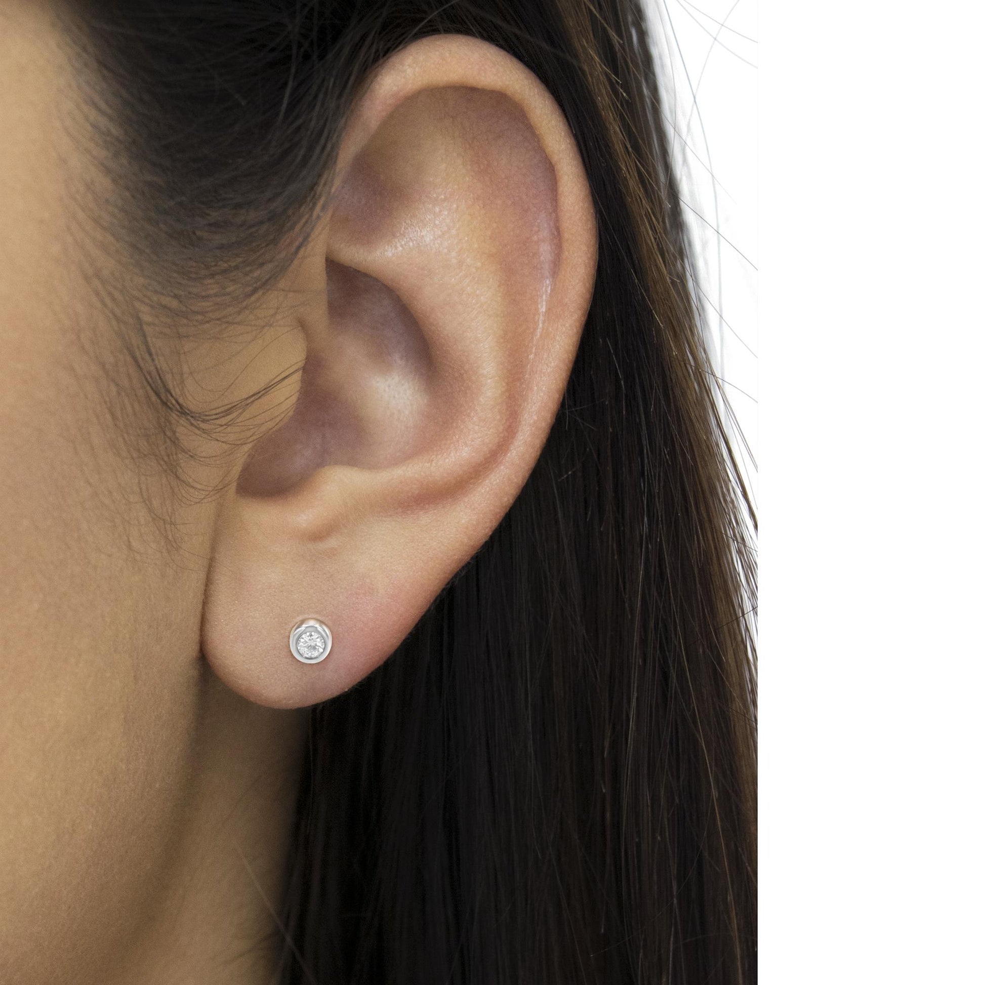 Close-up side view of a white metal stud earring with a round white gemstone in a bezel setting, worn on a model's ear.