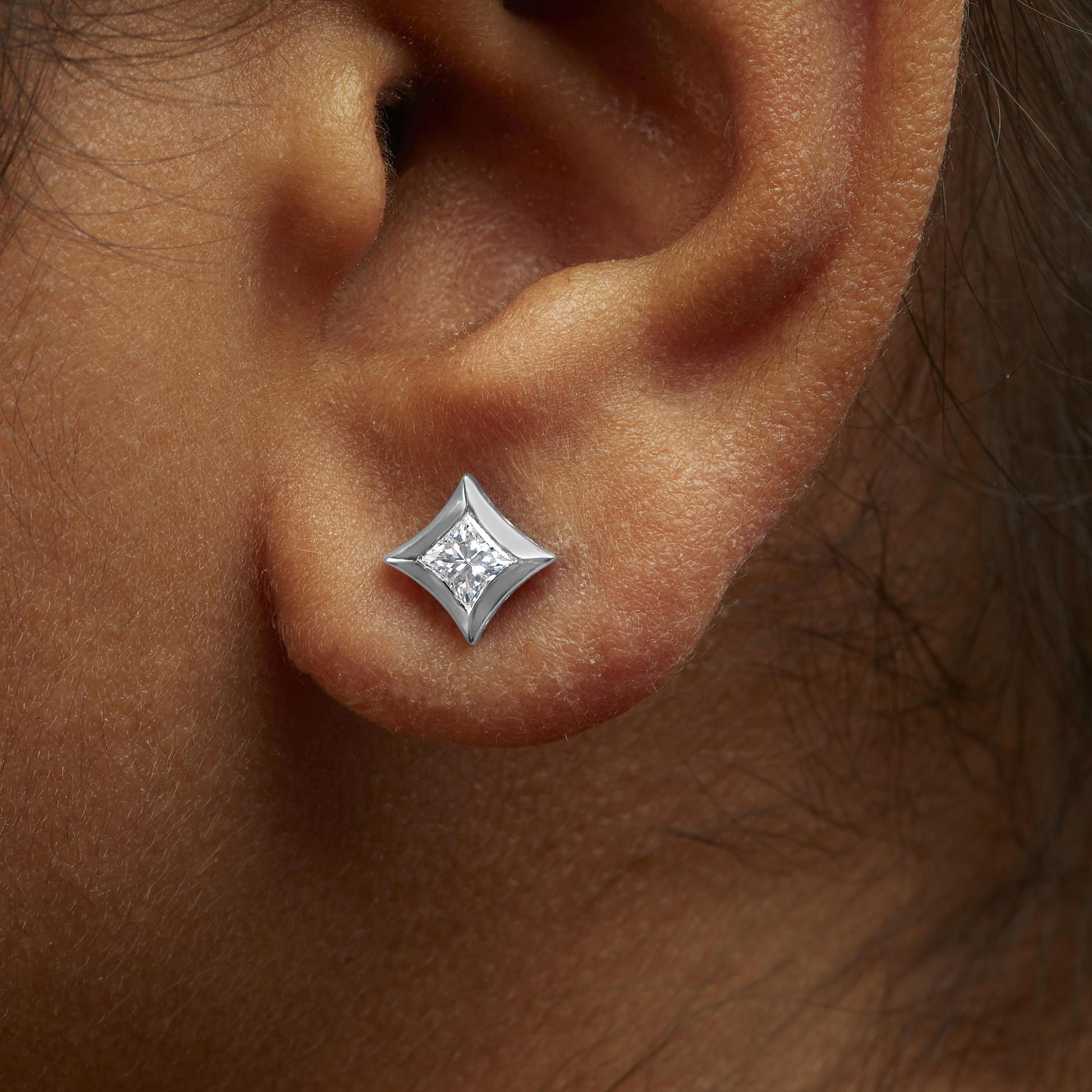 Close-up side angle of a white metal earring with a princess-shaped white gemstone in a bezel setting, worn on a model's ear.