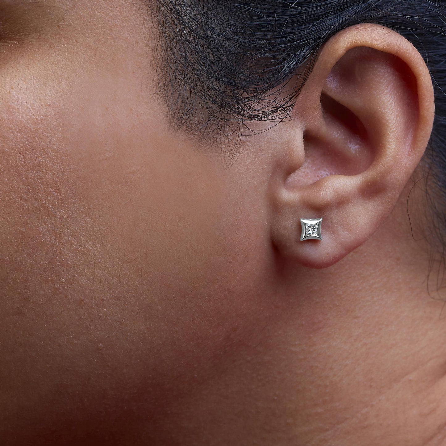 Close-up of a white metal princess shape stud earring with a white gemstone, worn on a model's ear, shot from the side.