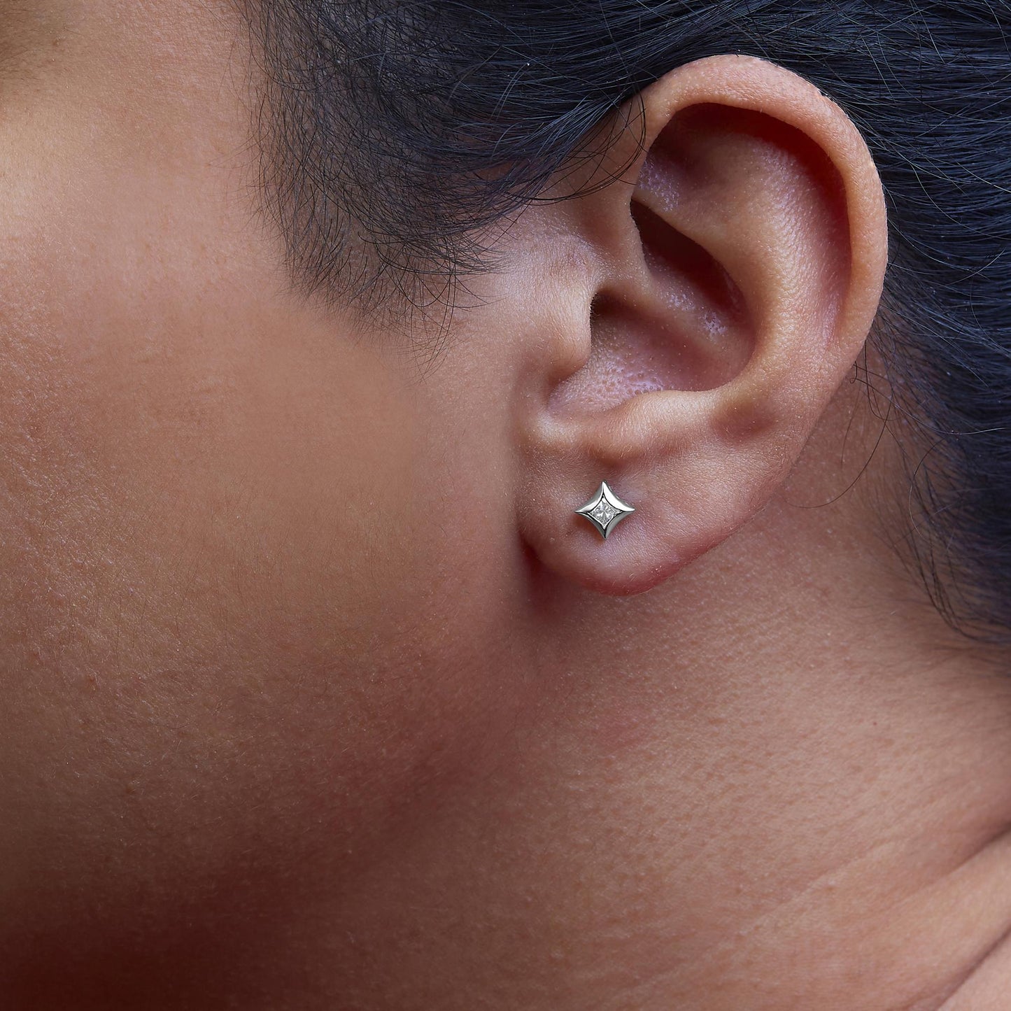 Close-up side view of an ear wearing a white metal stud earring with a princess-shaped white gemstone set invisibly.