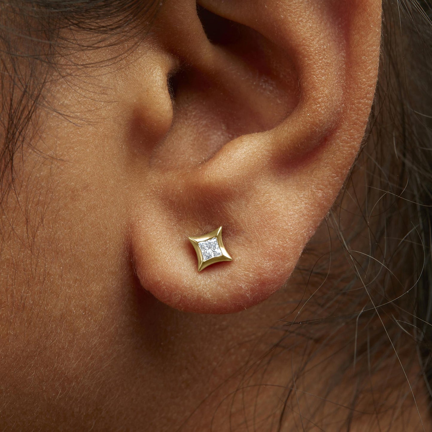 Close-up side view of an earlobe on a model wearing a yellow metal stud earring with a princess-shaped white gemstone set invisibly.