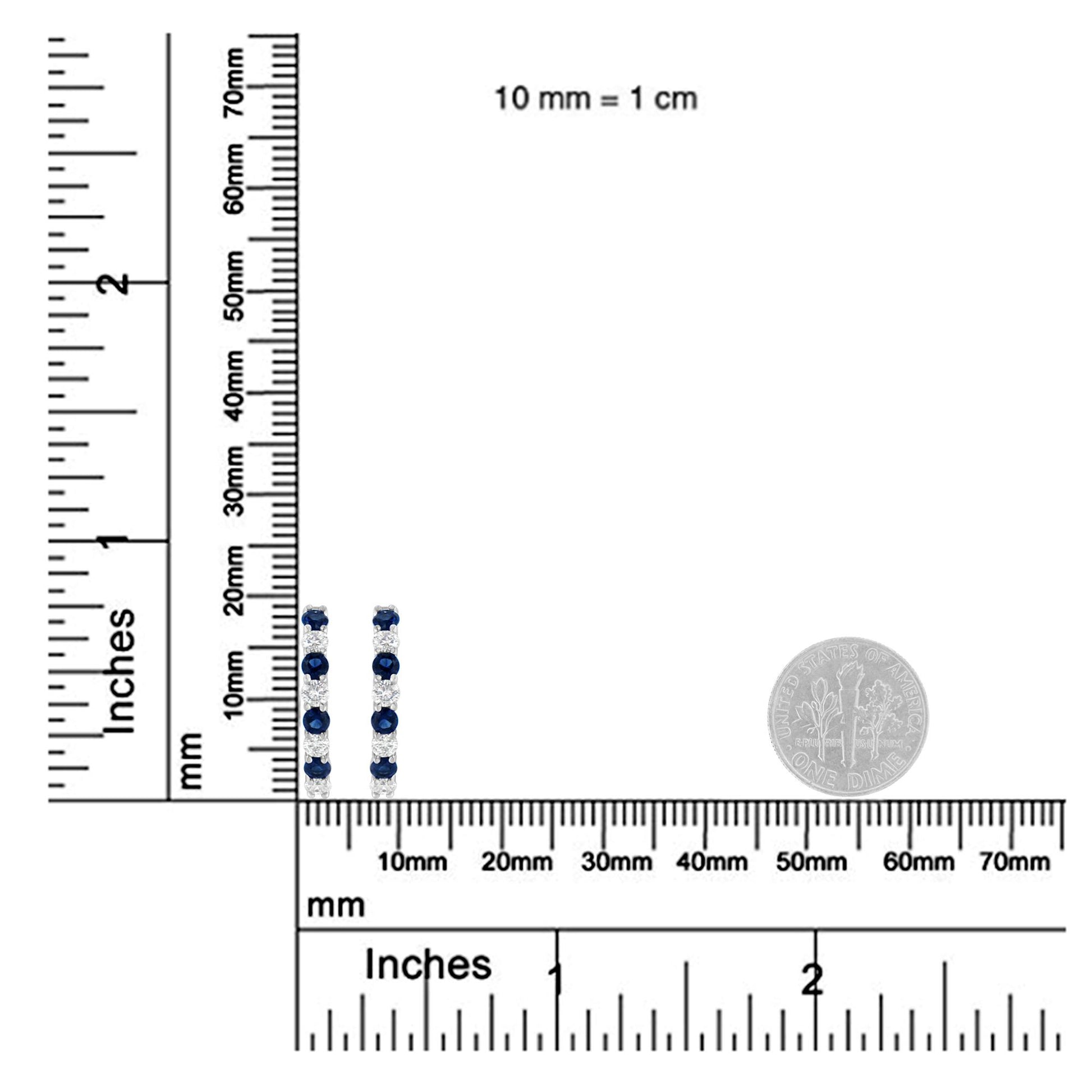 Pair of white metal earrings with alternating blue and white round gemstones in prong settings, shown upright on a ruler and scale for size reference with a coin beside them.