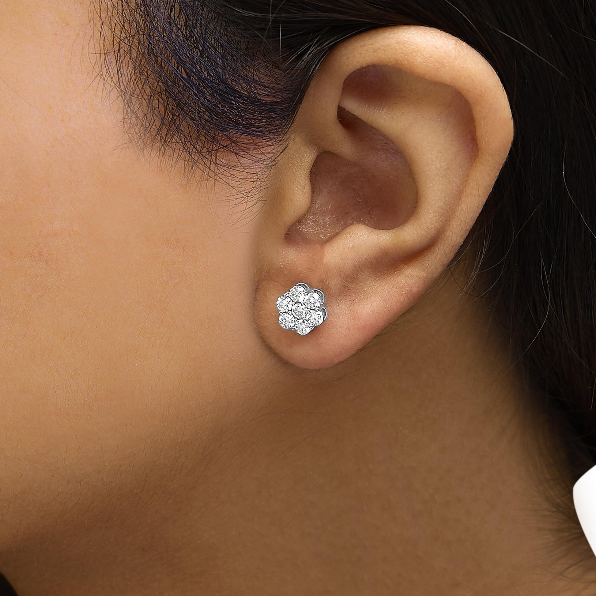 Close-up side view of a white metal flower-shaped stud earring with round white gemstones in prong settings, worn on a model's ear.