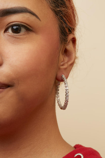 Close-up side view of a white metal hoop earring with prong-set round white gemstones, worn on a model.