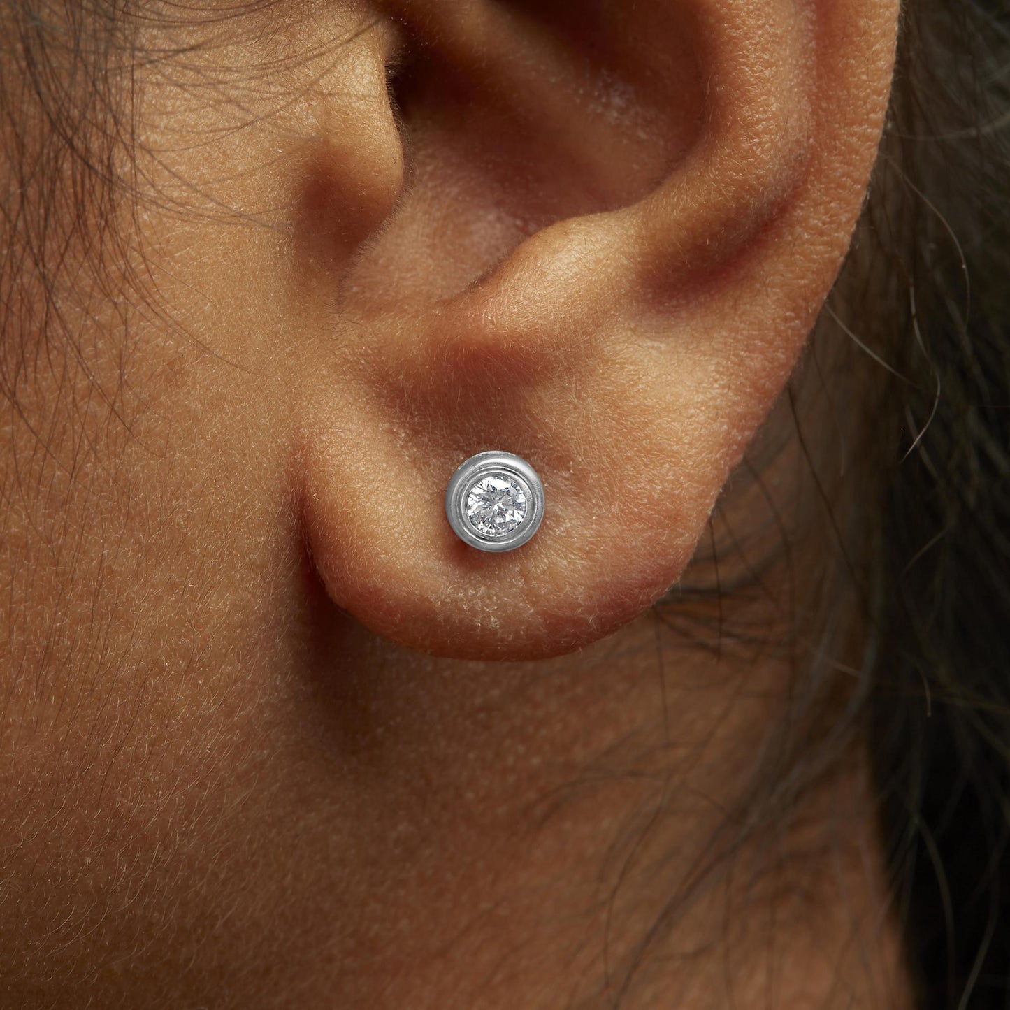 Close-up side view of a white metal stud earring with a round white gemstone in bezel setting, worn on a model's ear.