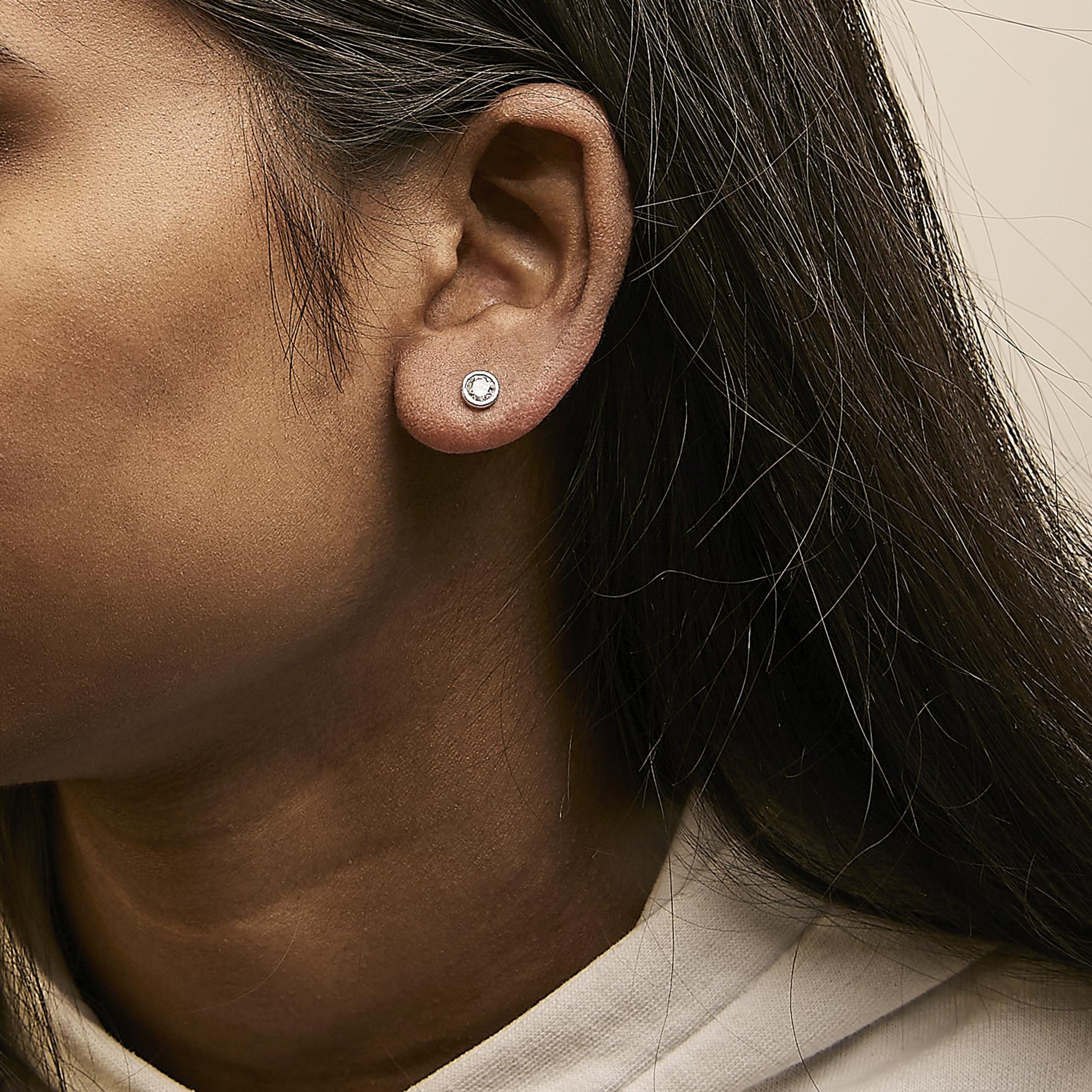 Close-up side view of a model wearing a white metal stud earring with a round white gemstone in a bezel setting.