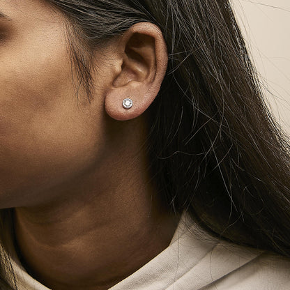 Close-up side view of a model wearing a white metal stud earring with a round white gemstone in a bezel setting.
