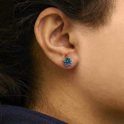 Close-up side profile of an ear wearing a yellow metal stud earring with a round blue gemstone in a four-prong setting.