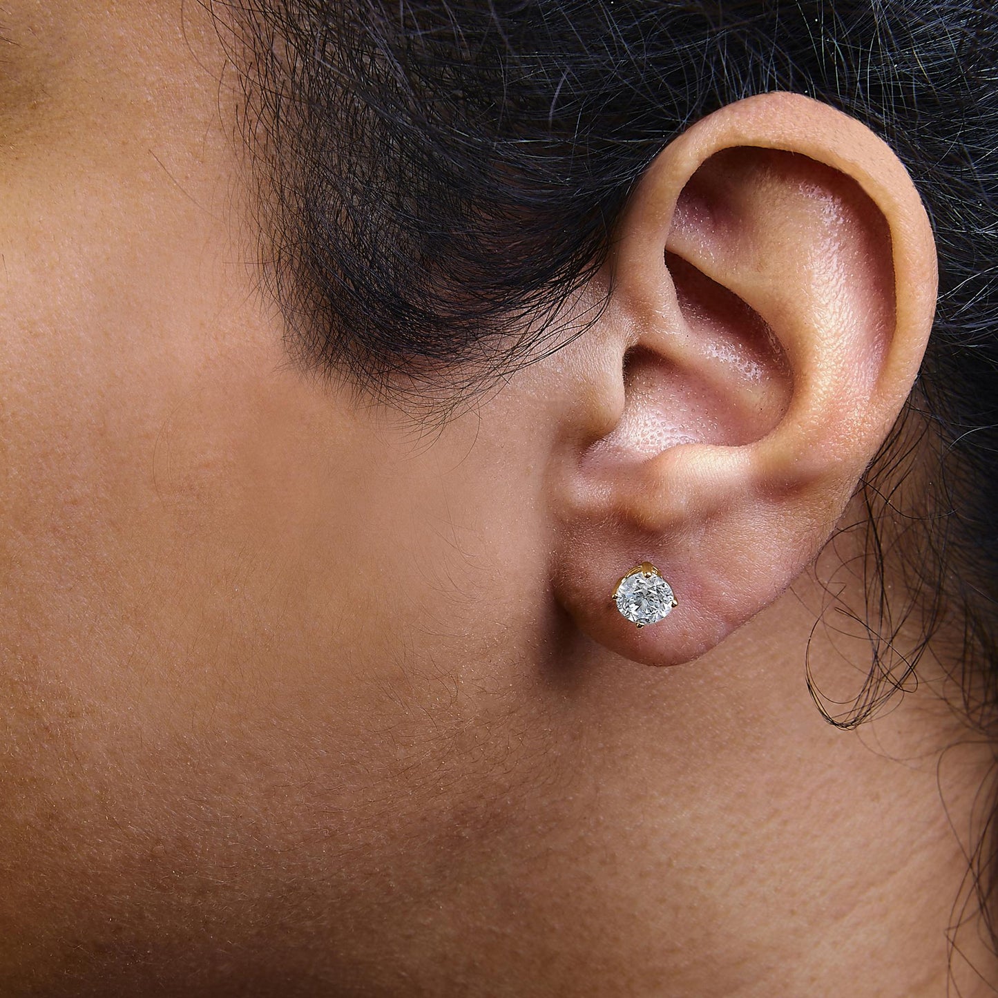 Close-up side view of a model wearing a yellow metal stud earring with a round white gemstone in a four-prong setting.