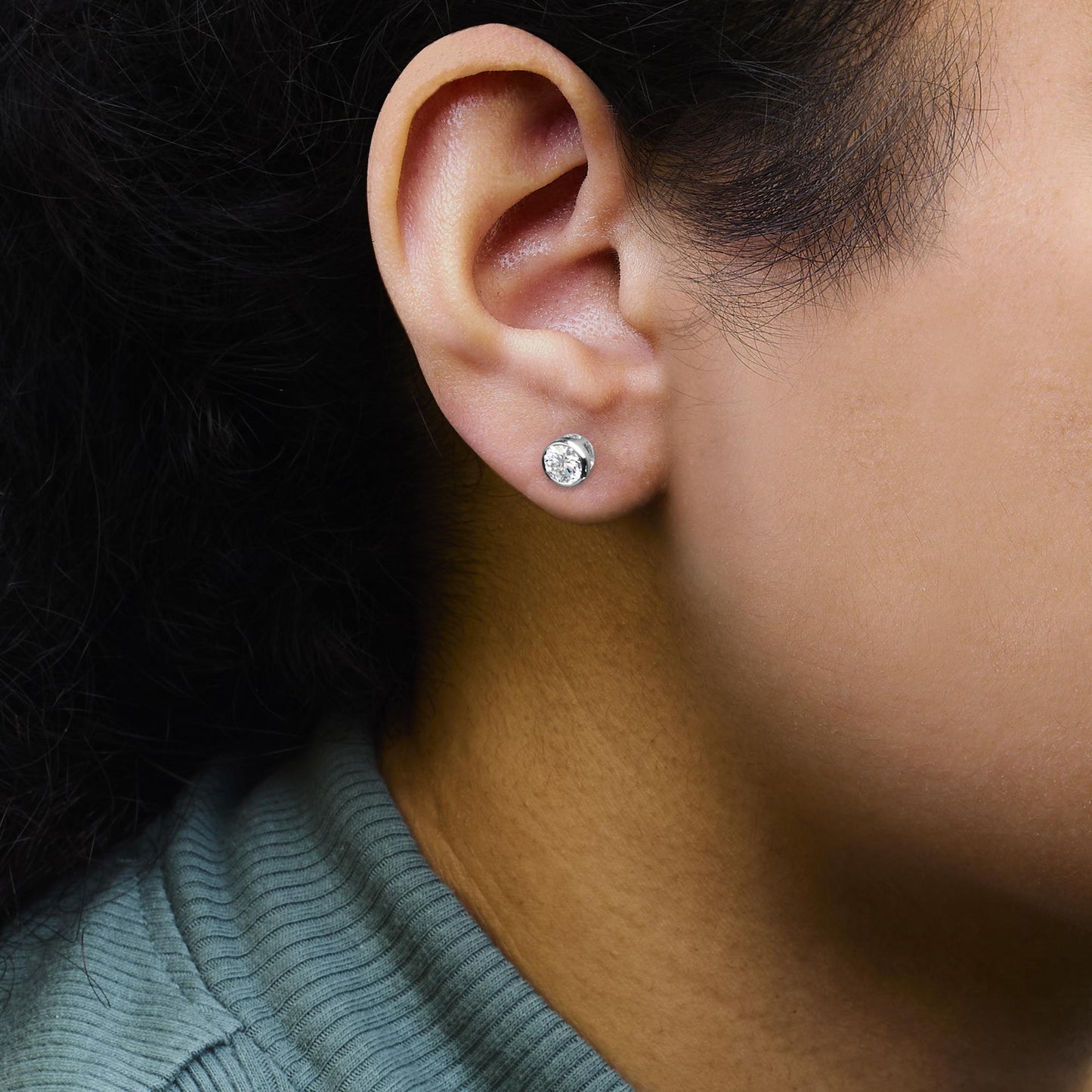 Close-up side view of a person wearing a white metal earring with a round white gemstone in bezel setting.