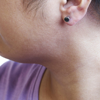 Close-up side view of a white metal stud earring with a round black gemstone in bezel setting, worn on a model's ear.
