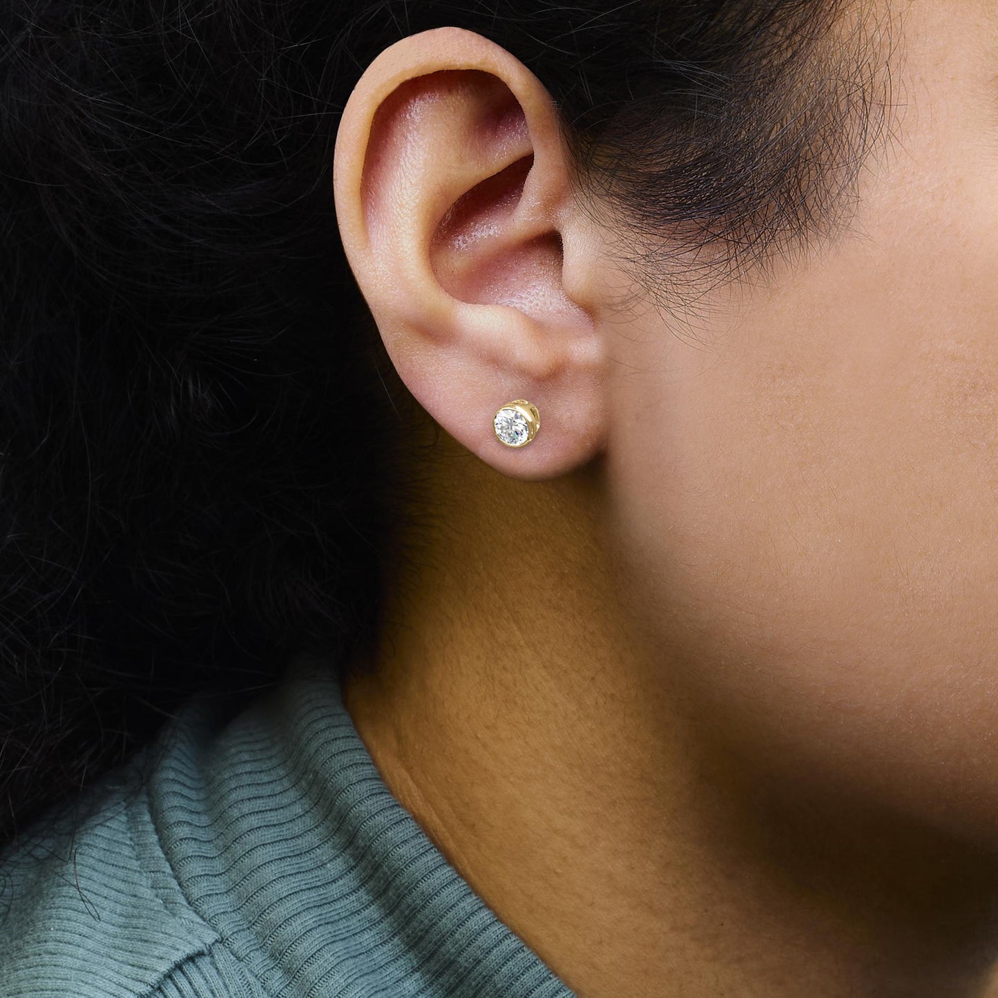 Close-up side view of a person's ear wearing a yellow metal stud earring with a round white gemstone set in a bezel setting.