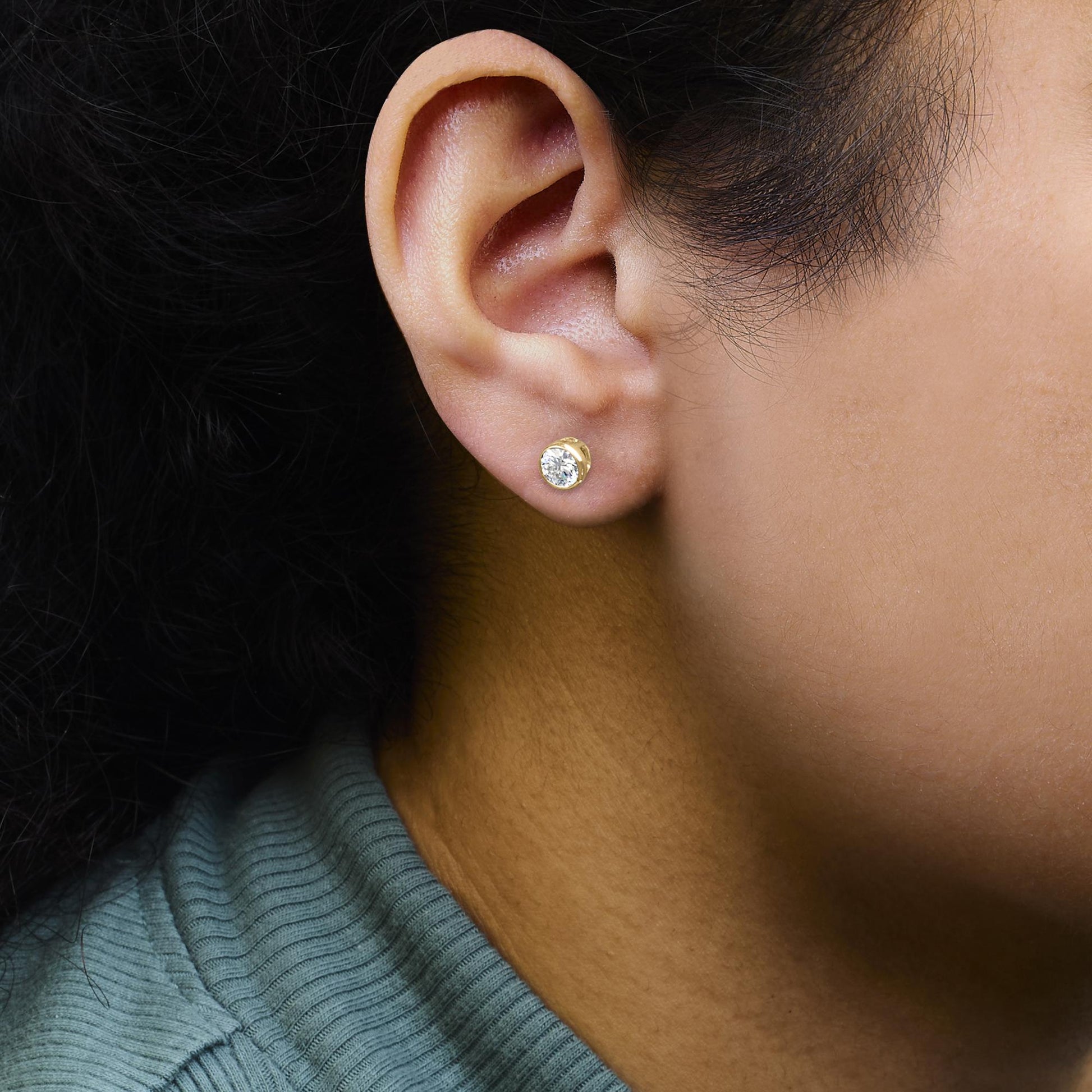 Close-up side view of a person's ear wearing a yellow metal stud earring with a round white gemstone set in a bezel setting.