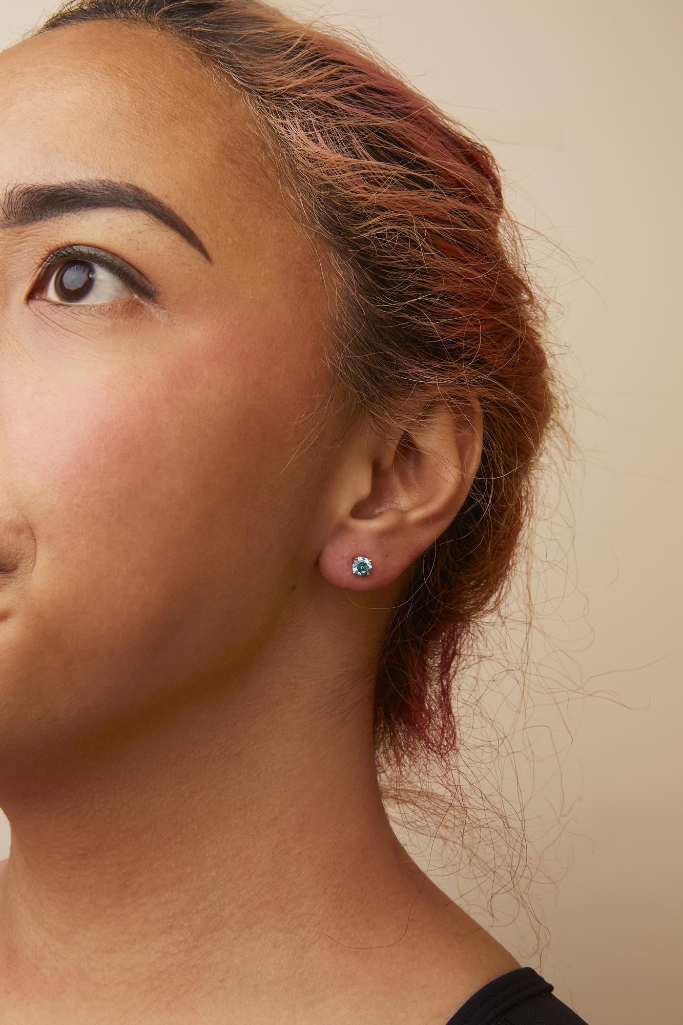 Close-up side view of a model wearing a yellow metal stud earring with a round aqua blue gemstone in a 4-prong setting.