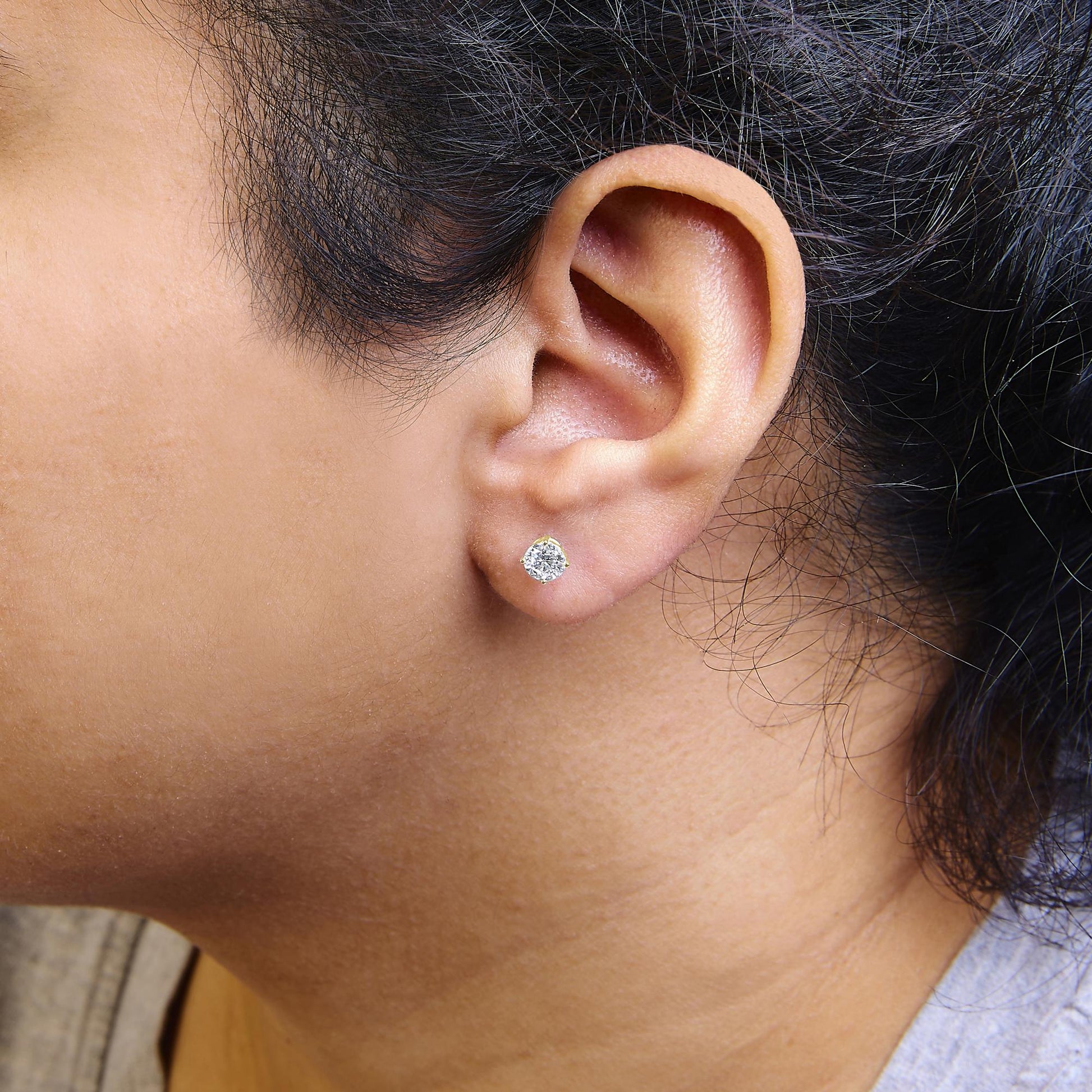 Close-up side view of a model wearing yellow metal stud earring with a round white gemstone in a 4-prong setting.