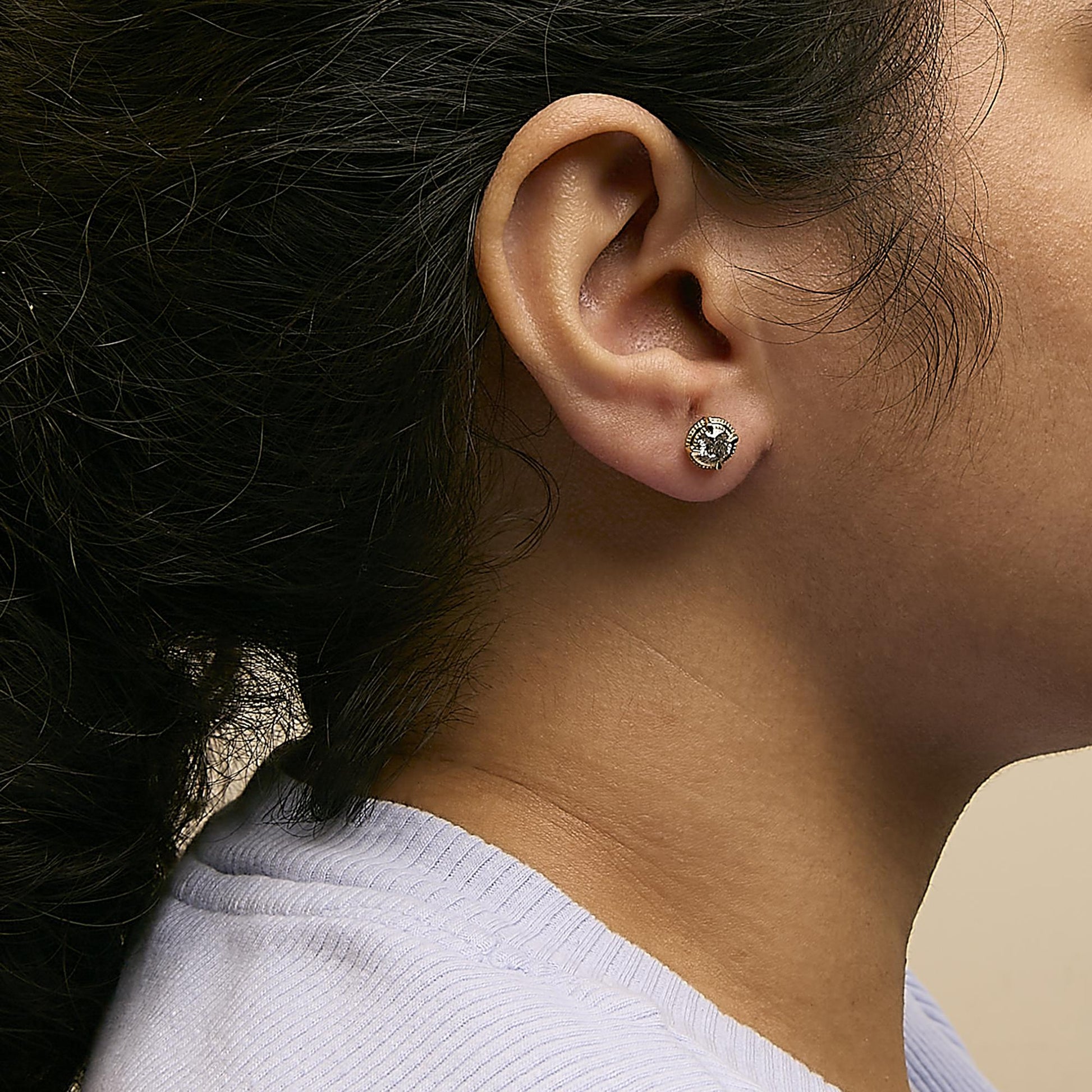 Close-up side view of a yellow metal stud earring with a round, brownish gemstone in a prong setting, worn on a model's ear.