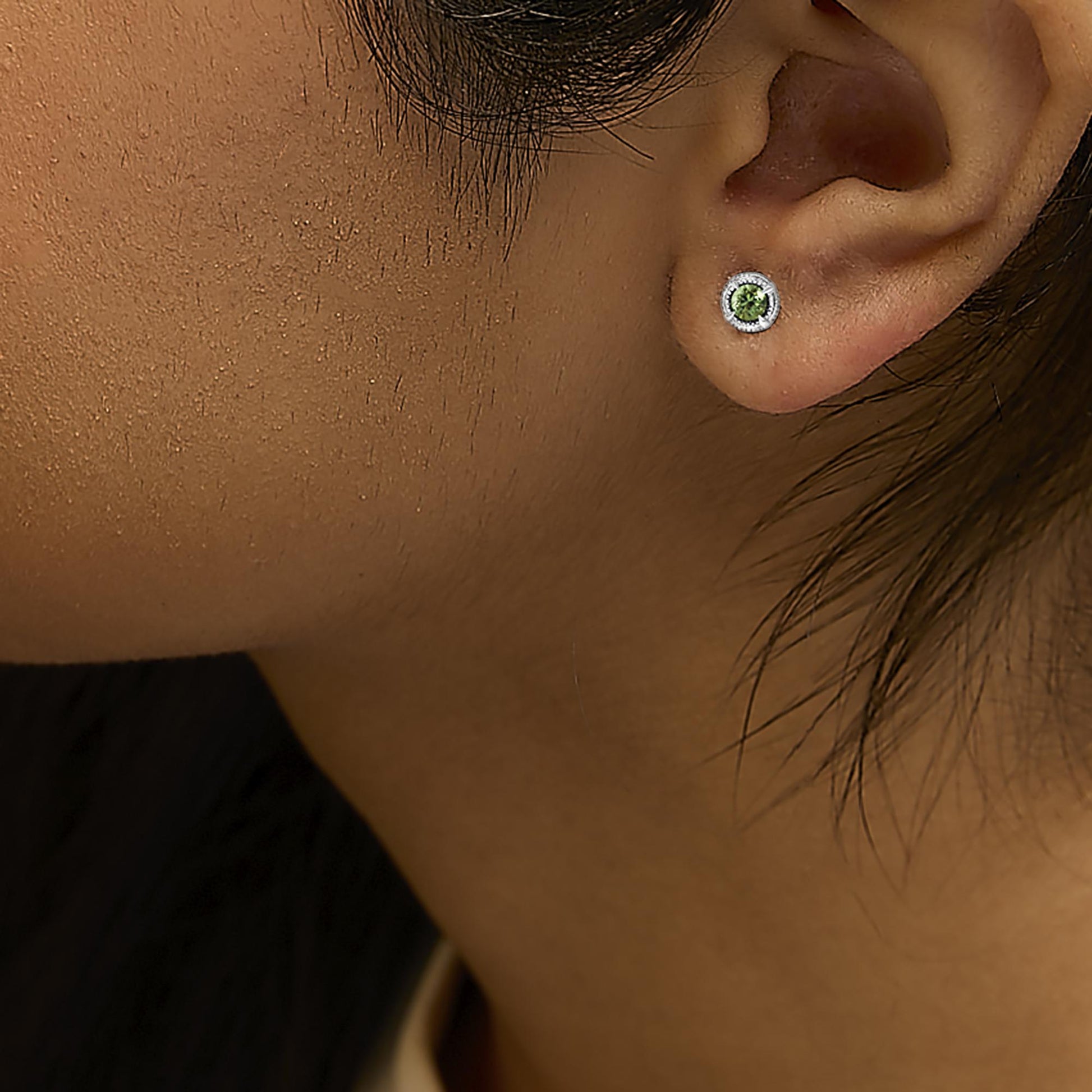 Close-up side view of a white metal stud earring with a round green gemstone in prong setting worn on a model's ear.